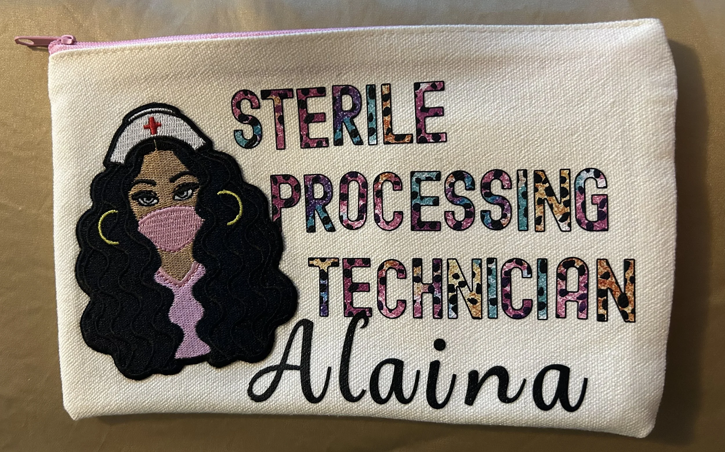 A personalized embroidered cloth bag with a cartoon nurse wearing a pink mask and white nurse hat. The bag has colorful, patterned text reading 'Sterile Processing Technician Alaina'.