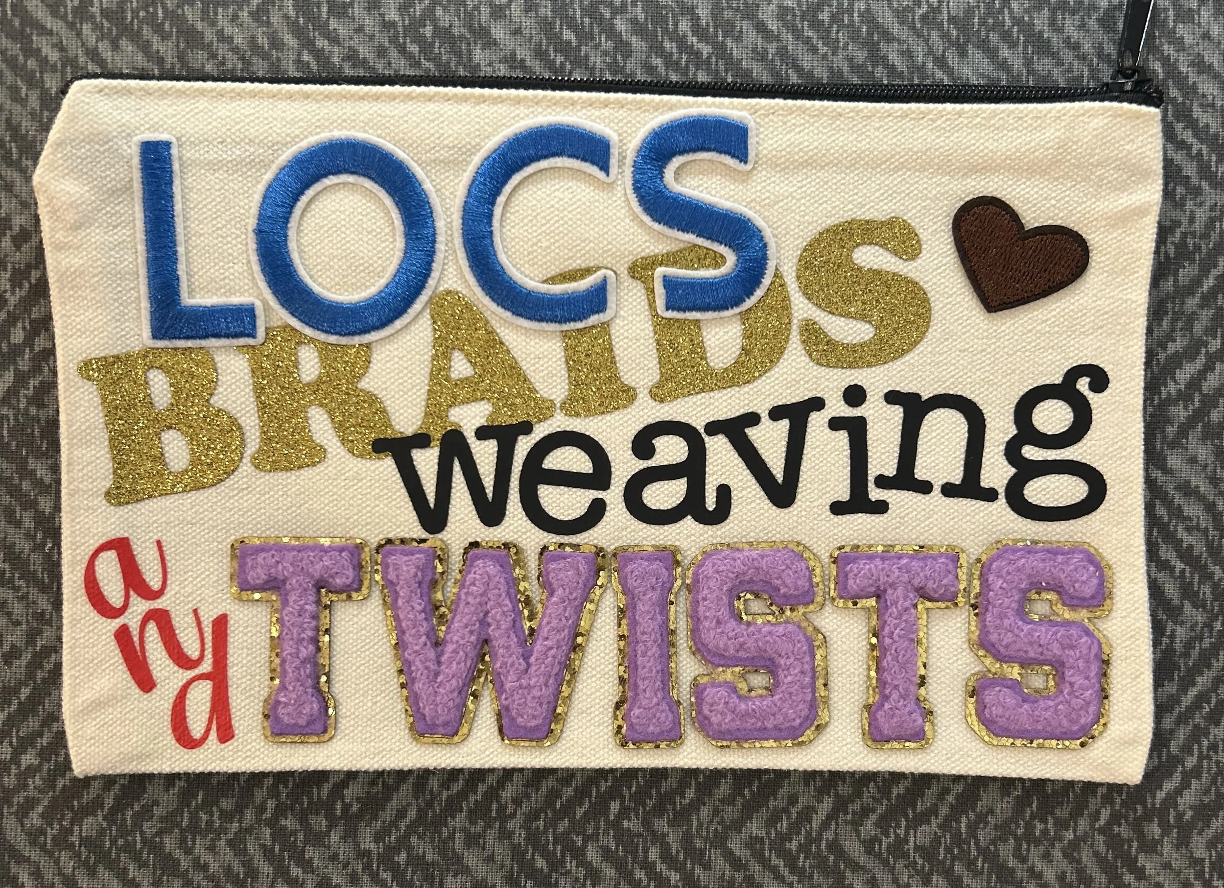 Decorative fabric panel with embroidered text reading 'LOCS BRADS & TWISTS' in colorful and glittery letters, along with a small brown heart design in the top right corner.
