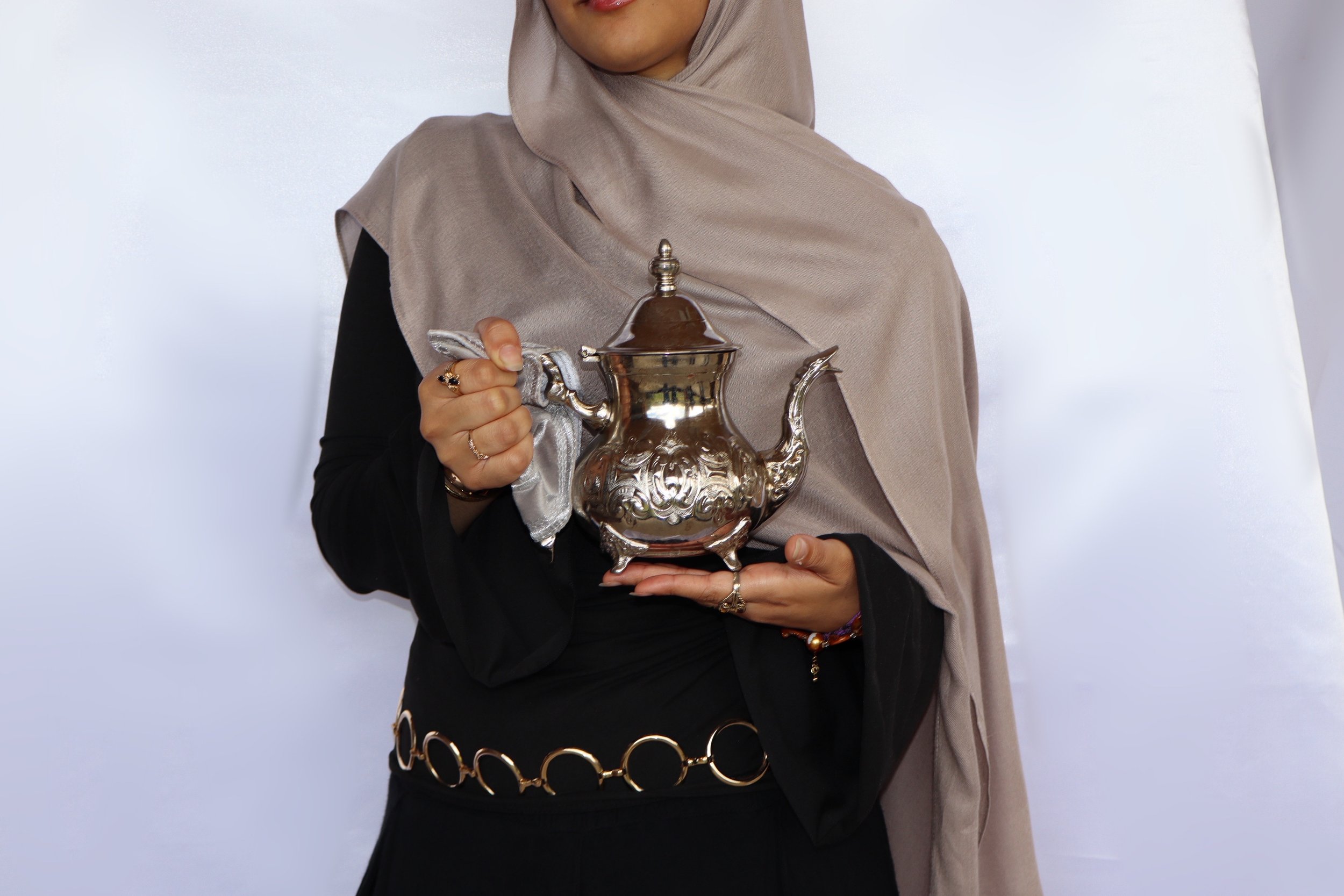 Person wearing a beige hijab and black dress holding a silver ornate teapot with a carved design, standing against a white backdrop.