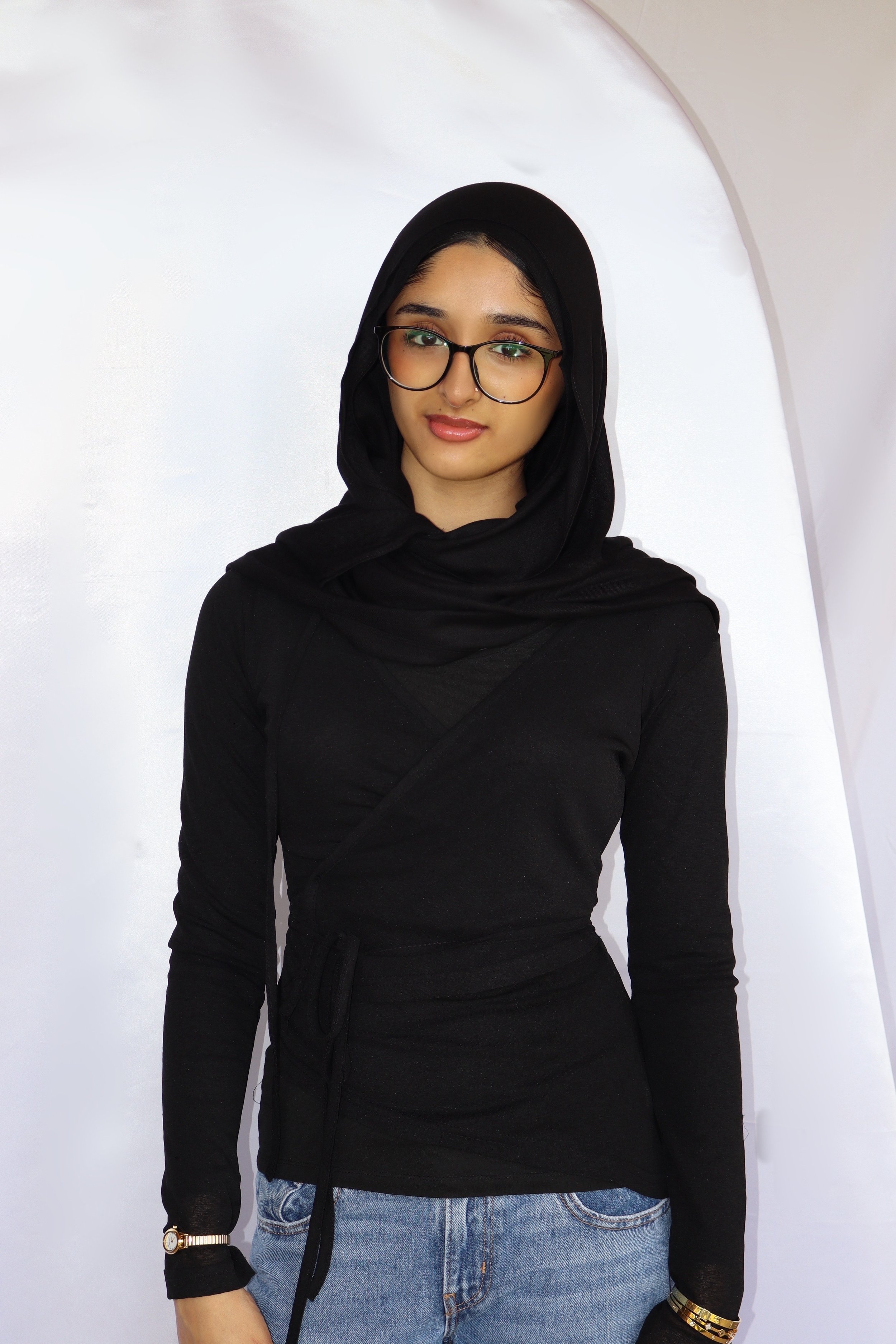 A young woman wearing a black hijab, glasses, a black top, and jeans, standing in front of a white backdrop.