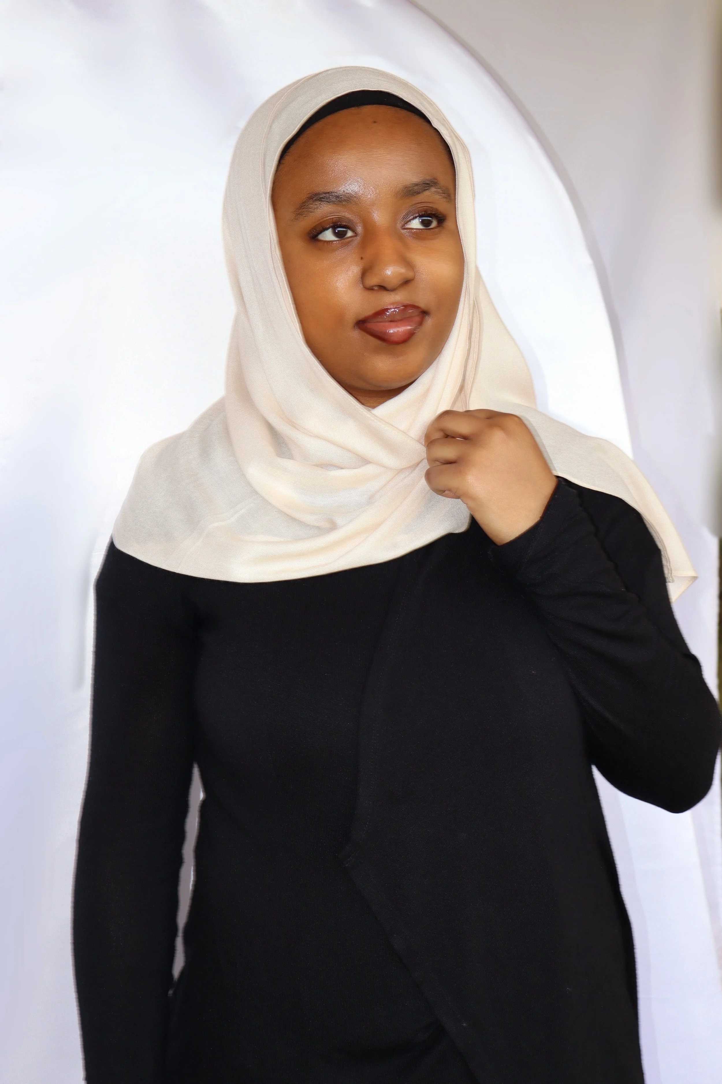 A woman wearing a beige hijab and black top, looking to her right with her hand on her chest, set against a white background.