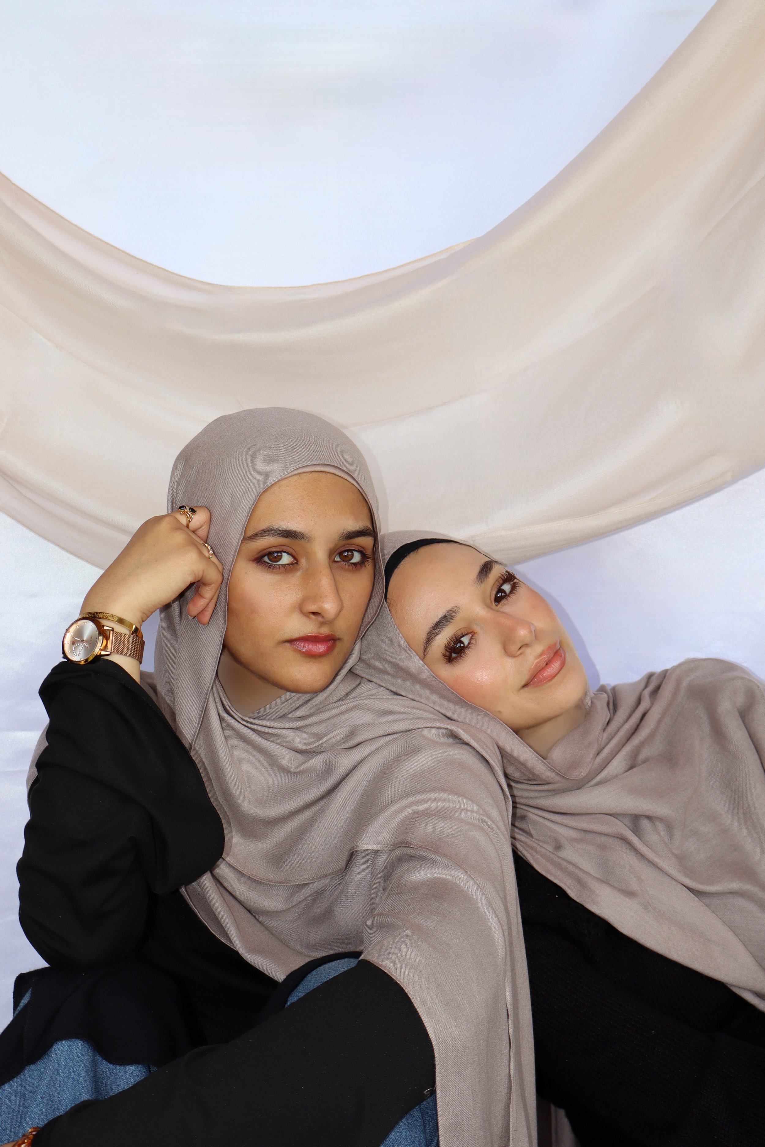 Two women wearing beige hijabs lying down, with one woman resting her head on the other woman's shoulder, against a light background with a draped fabric.