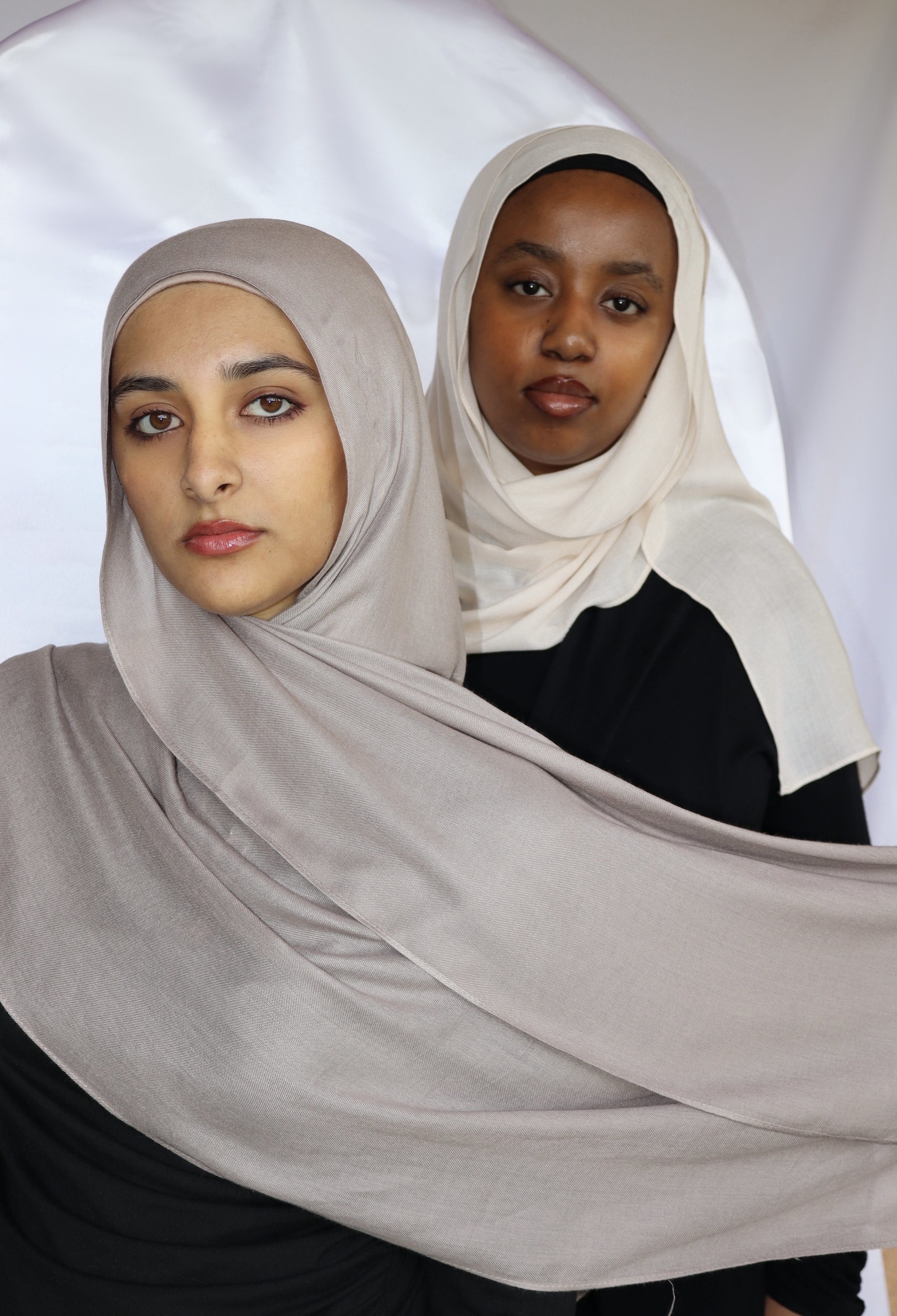 Two women wearing hijabs pose against a white backdrop, one in a light gray hijab and the other in a cream-colored hijab, both looking directly at the camera.