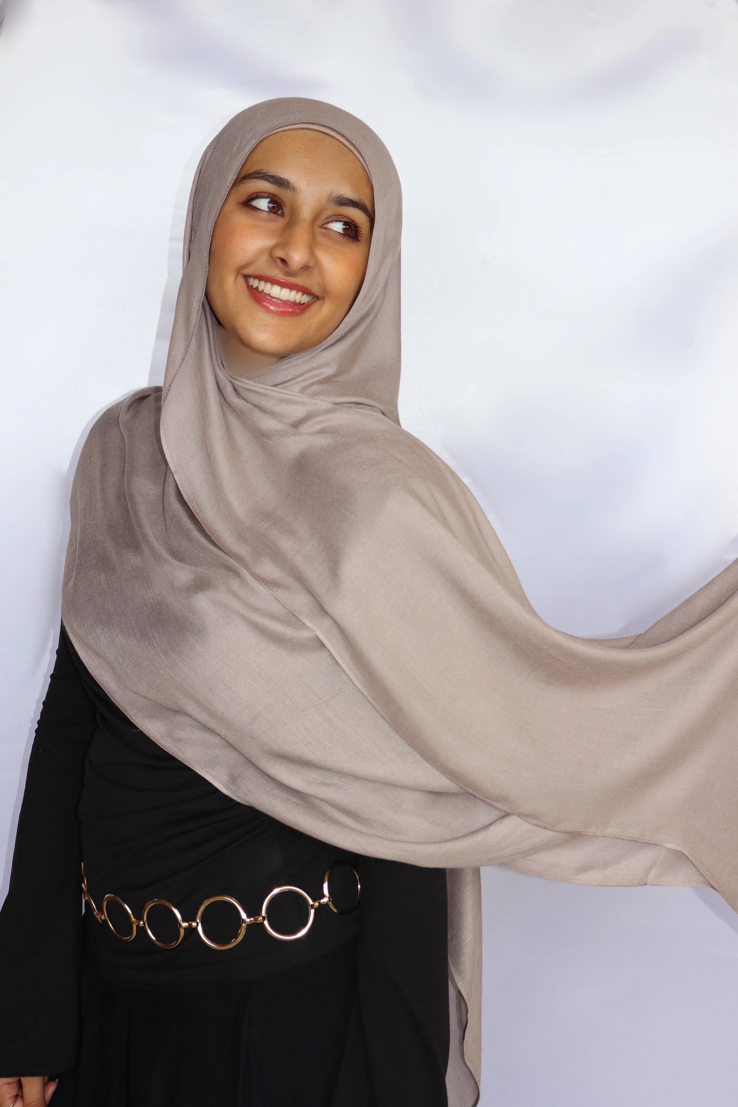 A woman with a beige hijab and black outfit smiling and looking to her left against a plain white background.