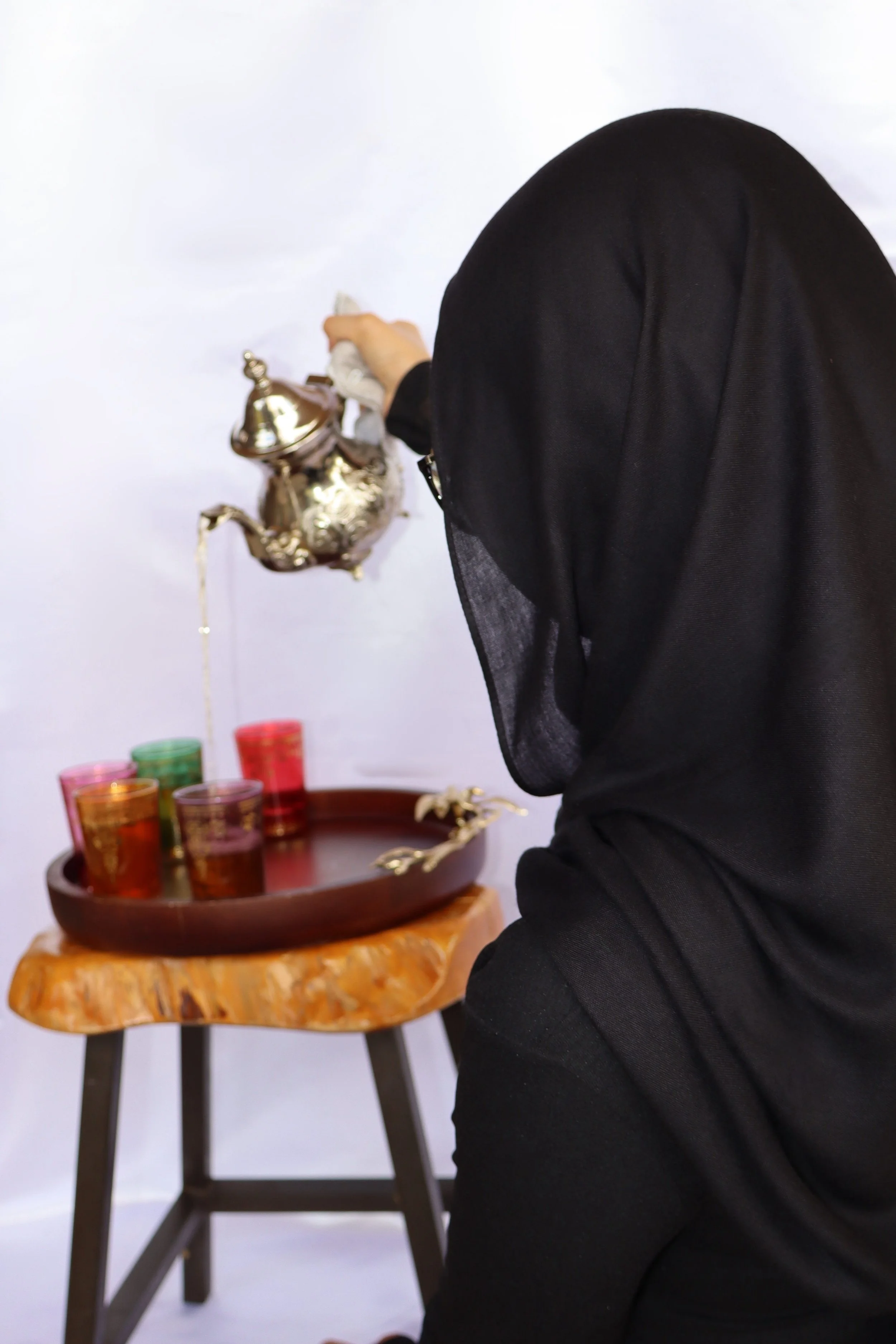 A woman in a black headscarf pouring tea from a decorative teapot into colorful glasses on a wooden table.