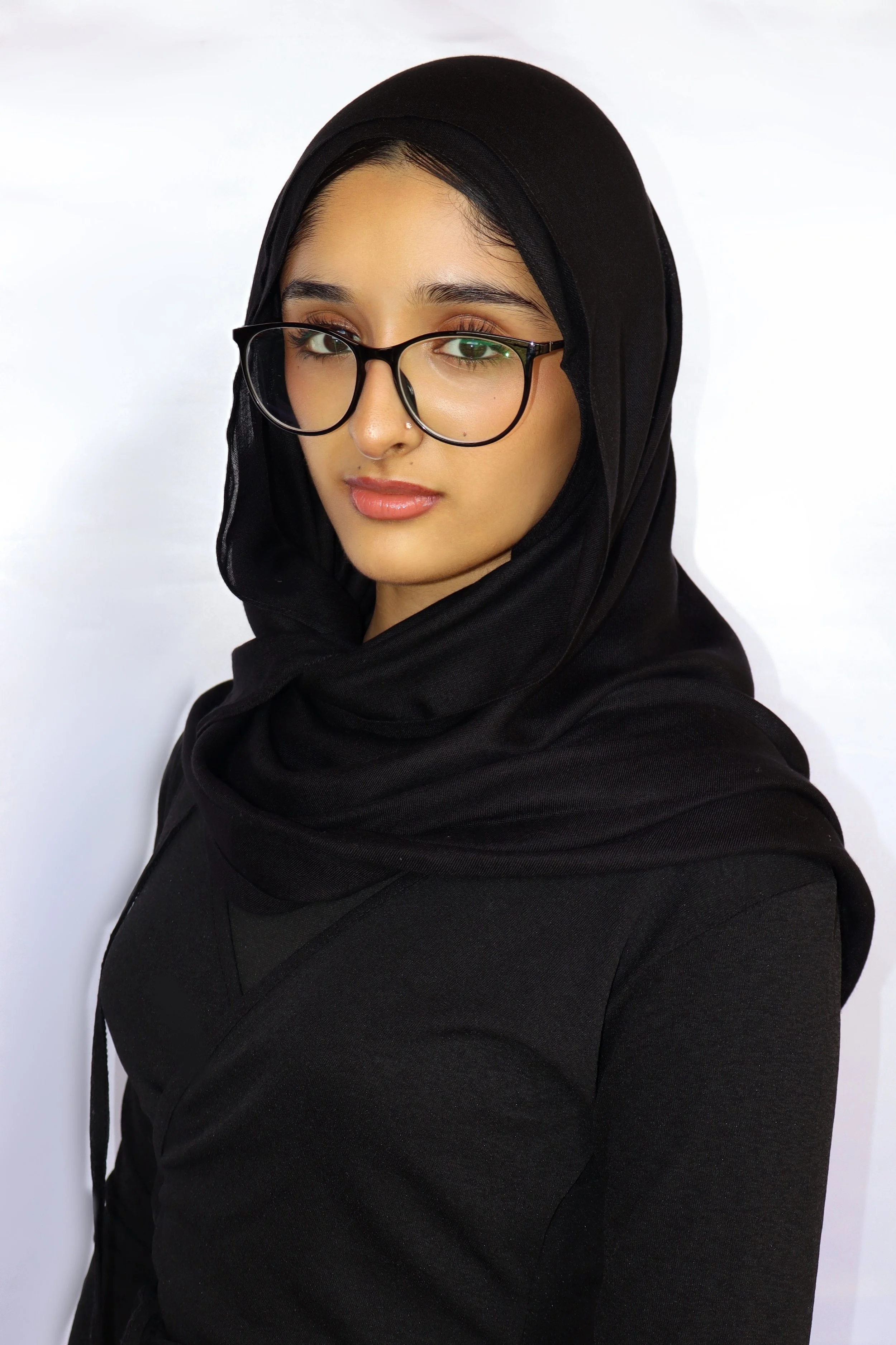 A young woman wearing a black hijab and glasses, looking at the camera with a slight smile, against a plain white background.