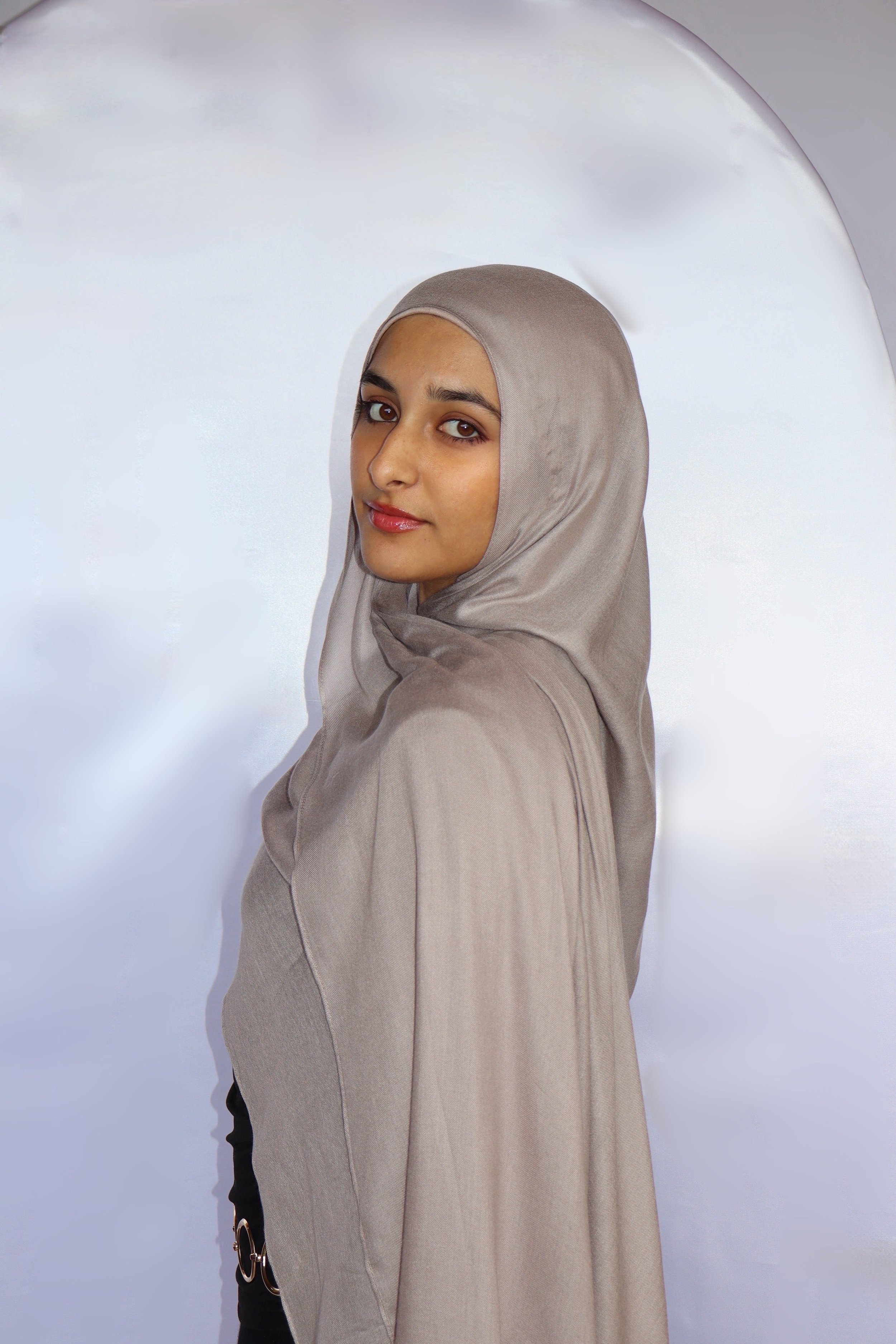 A woman in a beige hijab stands in front of a white backdrop, looking at the camera with a slight smile.