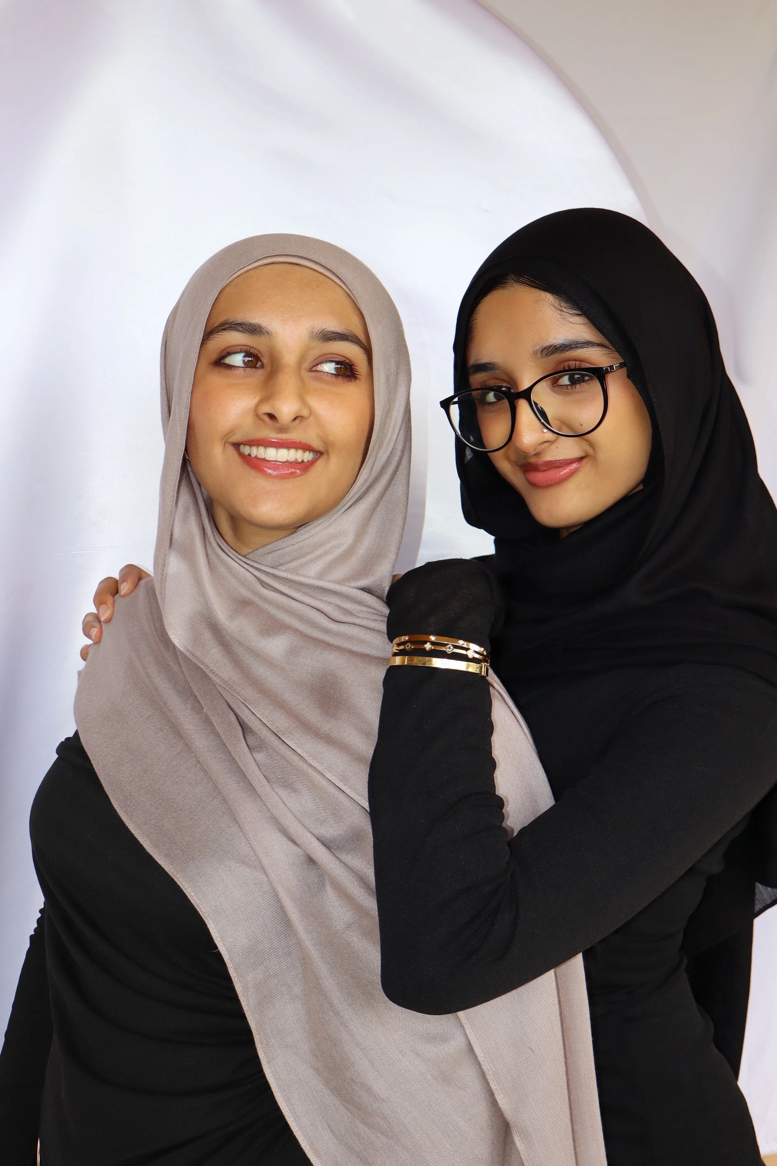 Two young women wearing hijabs smiling and posing together against a plain backdrop.
