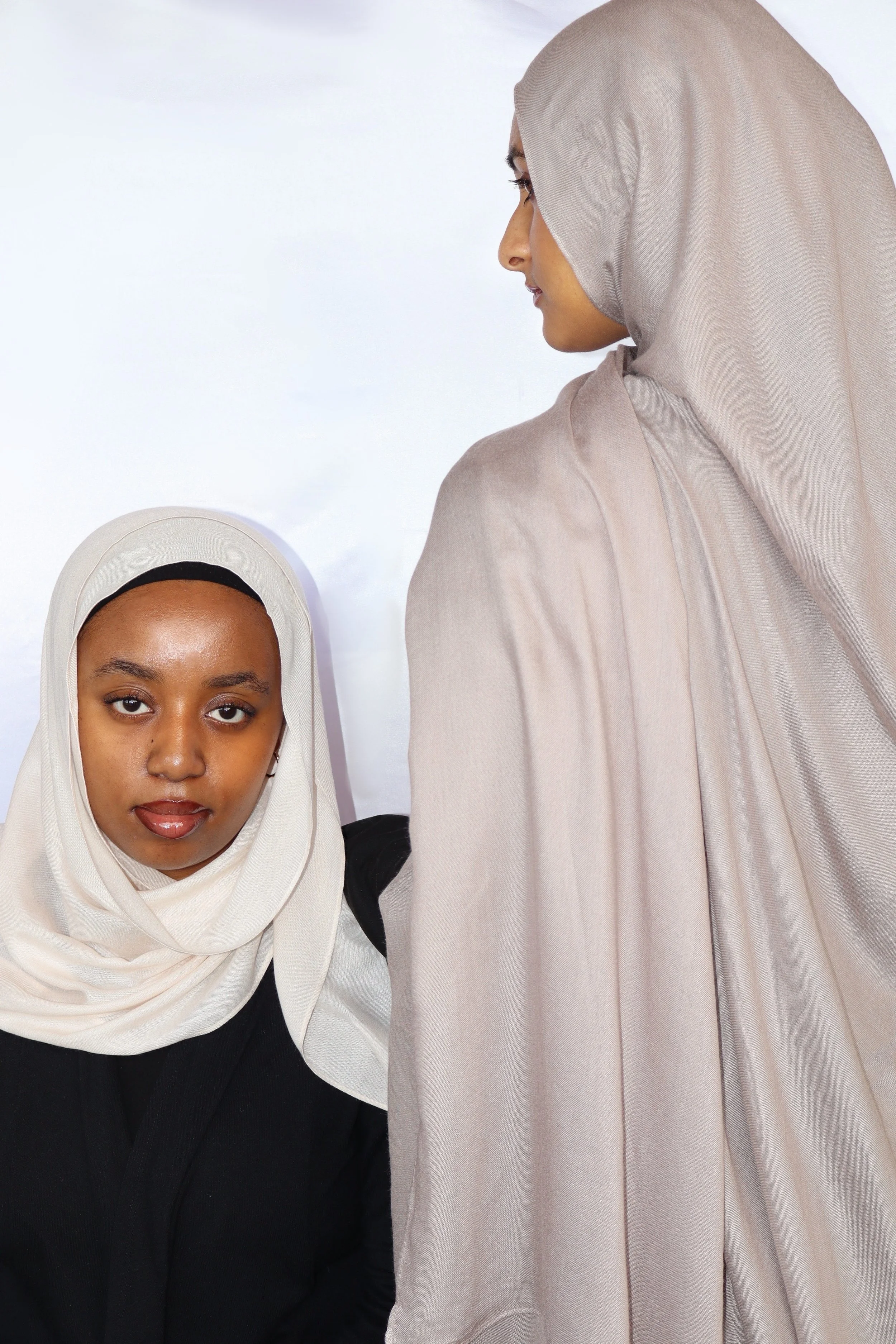 Two women in hijabs, one seated and one standing, against a plain white background.