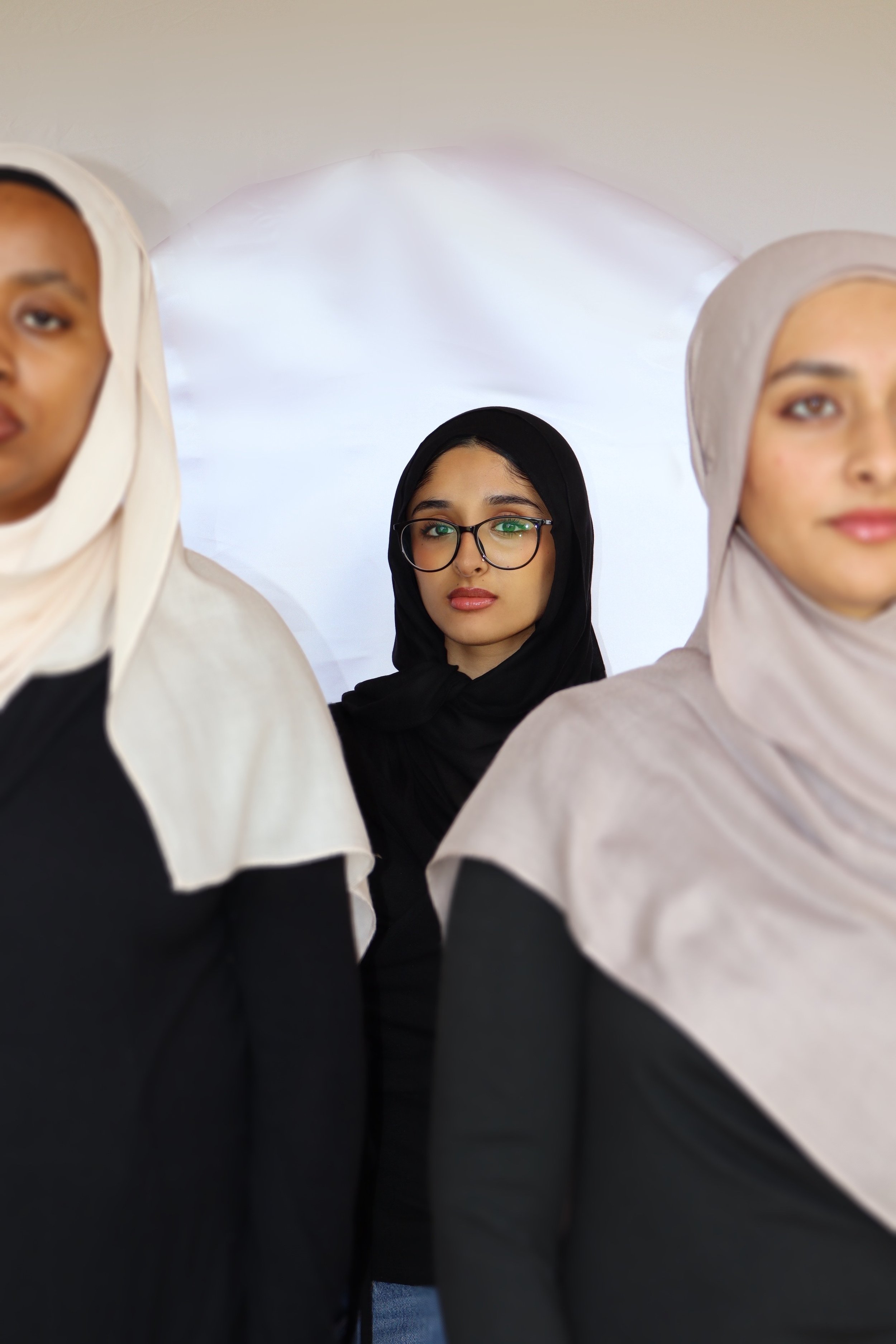 A group of women wearing hijabs, with one woman in the background wearing glasses and looking forward.