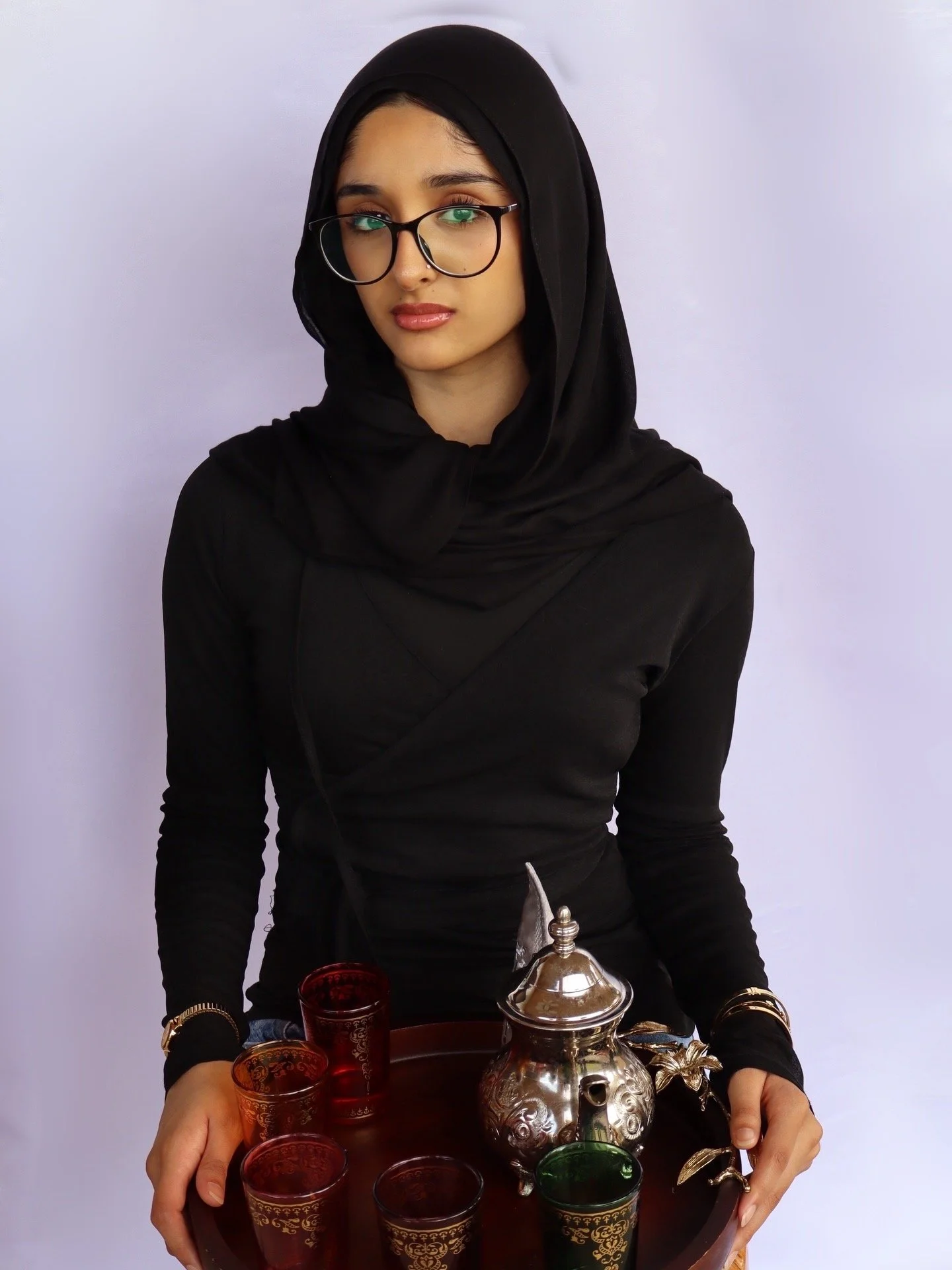 Young woman wearing glasses, a black hijab, and black clothing, holding a tray with Moroccan teapot and colorful teacups.