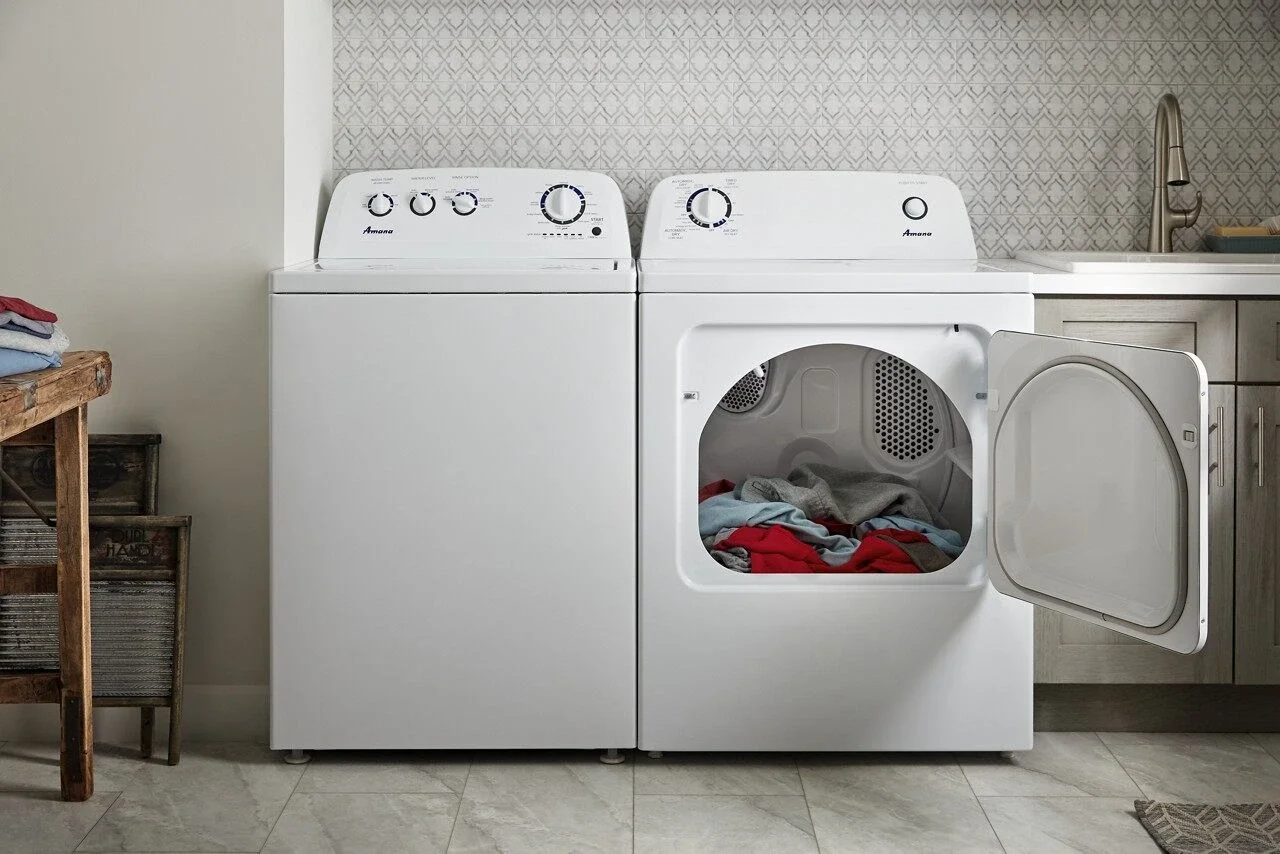 A laundry room with a white washing machine and dryer side by side. The dryer door is open, showing clothes inside. There is a wooden table with folded clothes to the left and a sink with a faucet and a cabinet to the right.