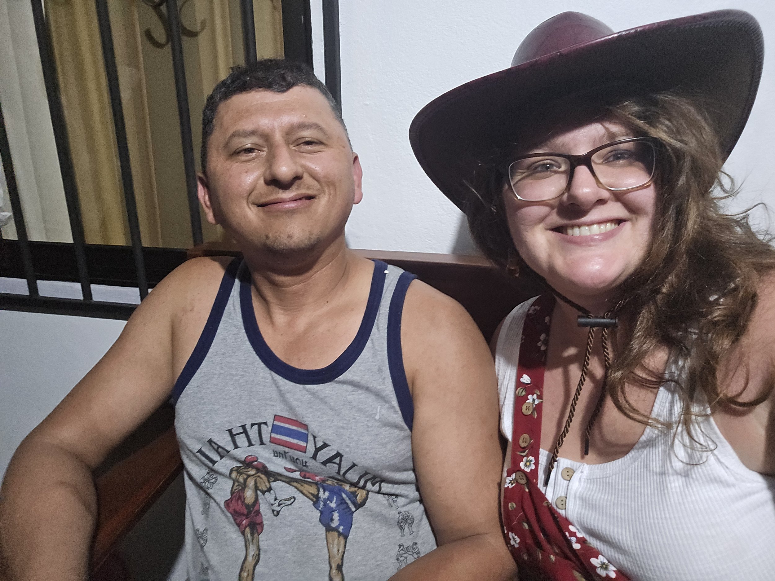 A man and a woman smiling for a photo, sitting closely together. The man is wearing a sleeveless tank top with a graphic of two basketball players. The woman is wearing glasses, a wide-brimmed hat, and a red bandana with white floral prints around her neck.