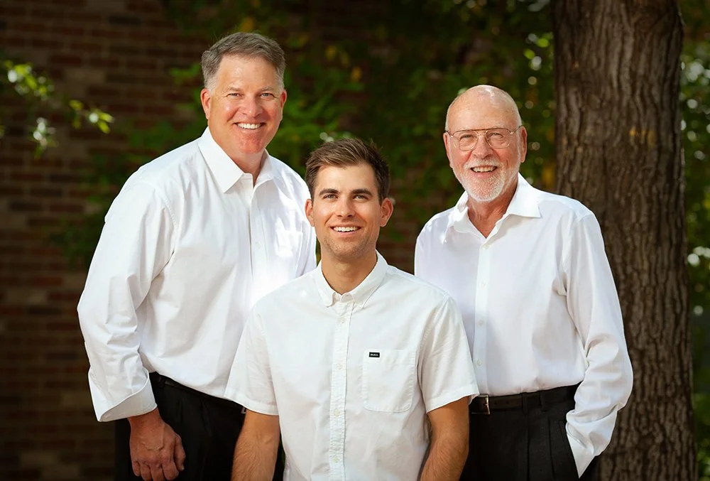 Three men standing outdoors in front of a tree, all dressed in white shirts and smiling.