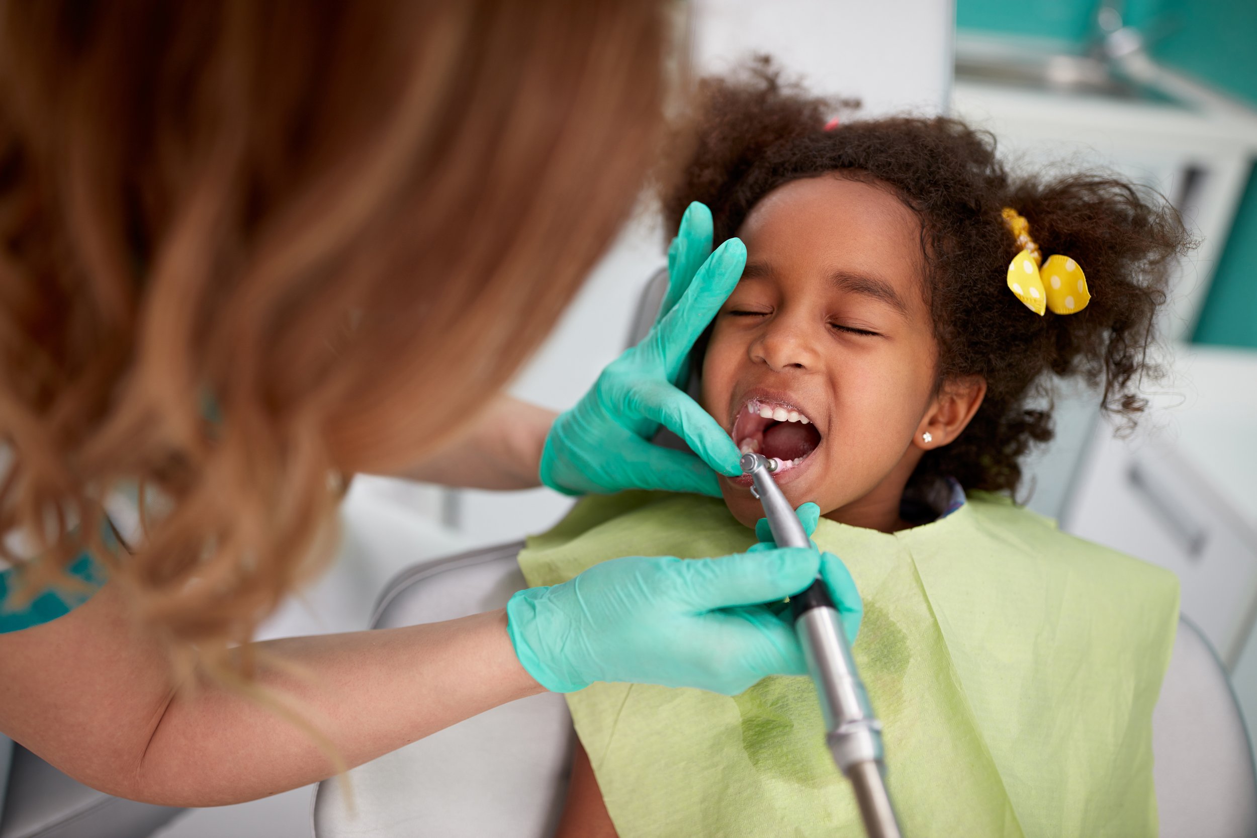 A young girl with curly hair and a yellow bow getting dental care from a dental professional wearing teal gloves.