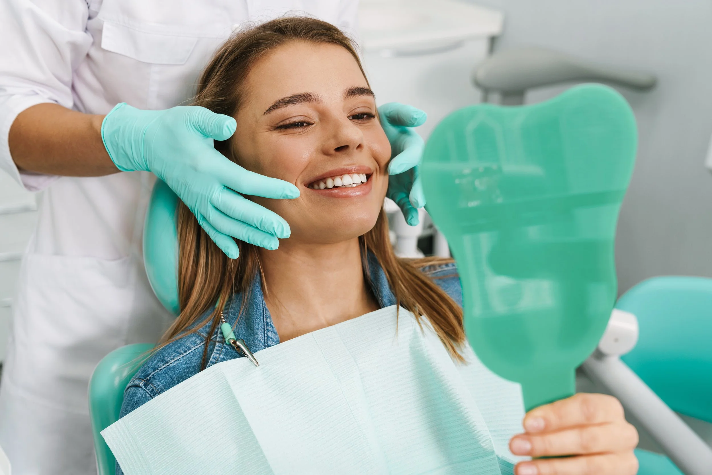 A woman sitting in a dental chair, smiling and looking at a mirror, during a dental appointment. A dental professional wearing gloves is adjusting her face.