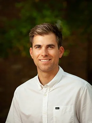 A man with gray hair smiling outdoors, wearing a white button-up shirt.