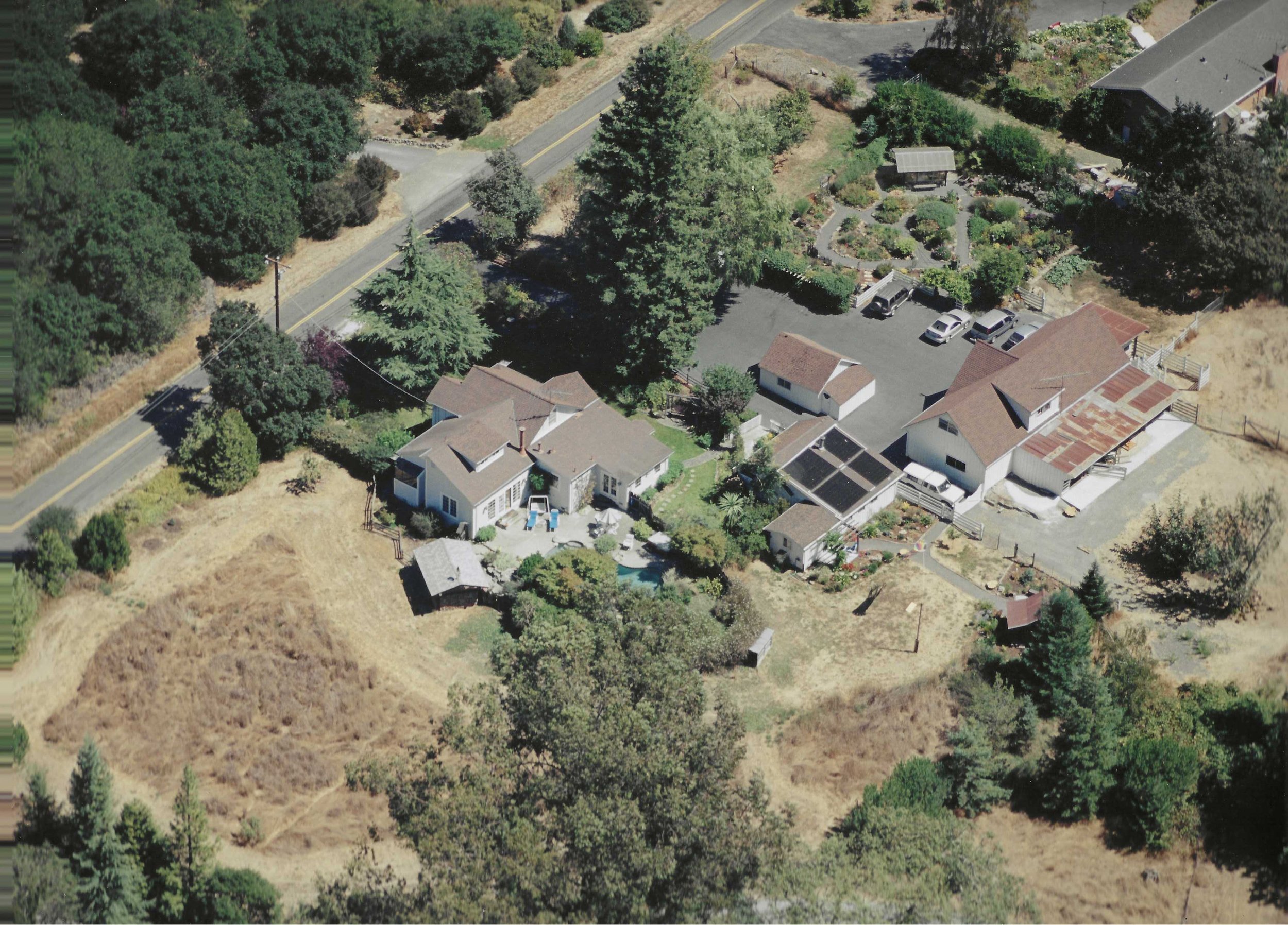 An aerial view of a residential neighborhood with several houses, trees, a driveway, and a parking area, surrounded by a dry hillside and a road.