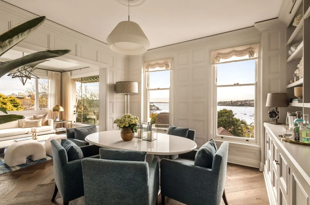 Bright living and dining area with large windows overlooking water and boats, white walls, modern decor, and a round dining table with six chairs.