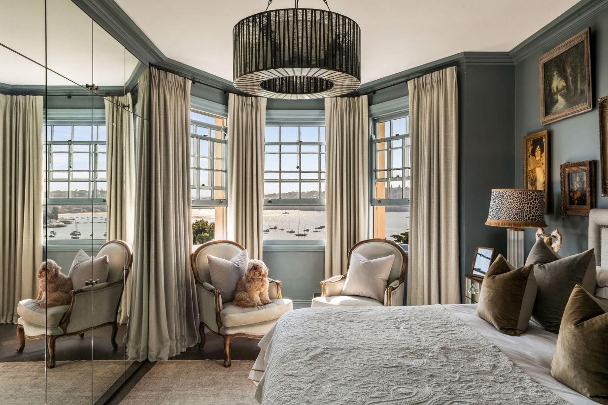 A cozy bedroom with a view of a waterscape through three large bay windows, decorated with light-colored curtains, a bed with multiple pillows, a nightstand with a lamp, two upholstered armchairs with small dogs sitting on the cushions, a black circu
