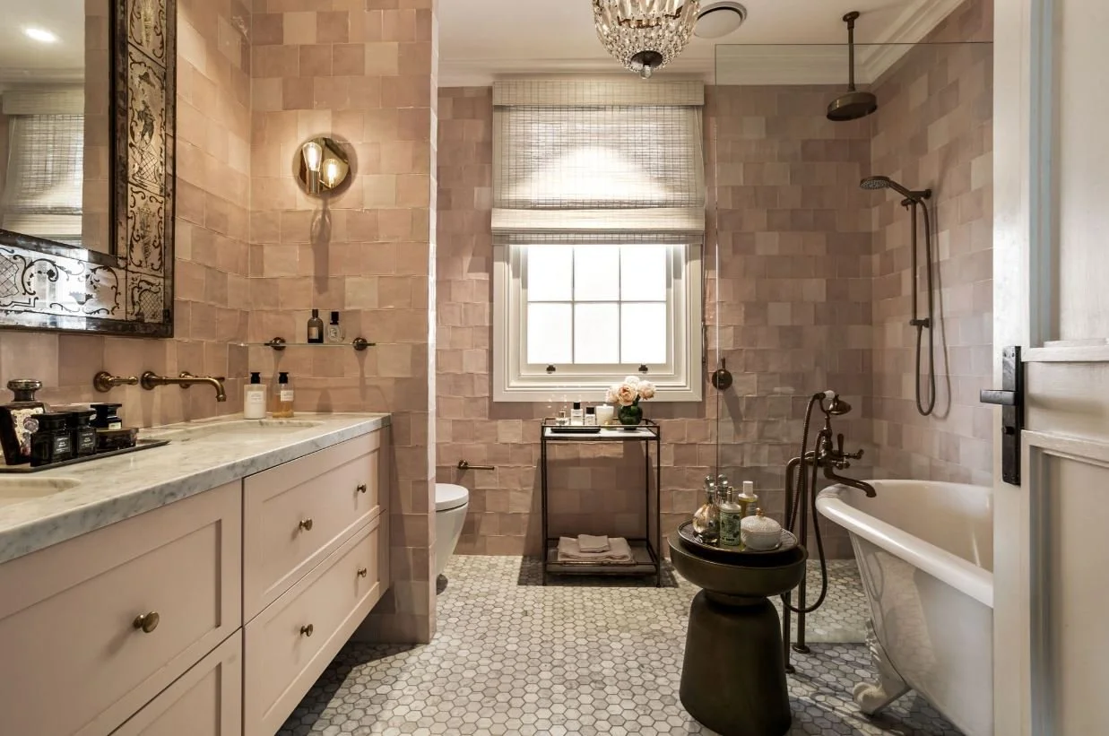A spacious bathroom with pink tiled walls and white hexagon tile flooring. Features a white vanity with a marble countertop, a large mirror, a stone bathtub, and a shower area with a rainfall showerhead. Contains a small black table with towels and d