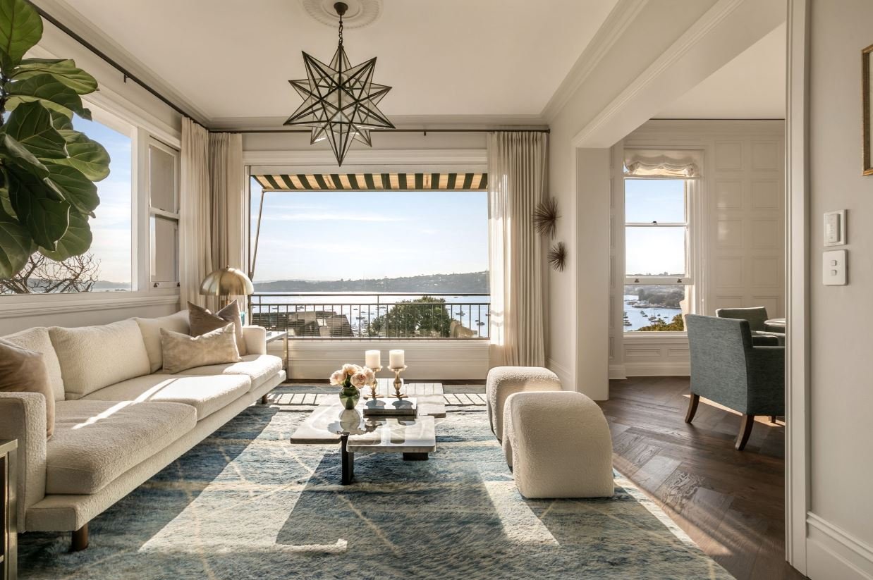 Living room with large windows showing a river view, white couch with beige and brown pillows, coffee table with candles and flowers, rug, lamps, and decorative wall art.