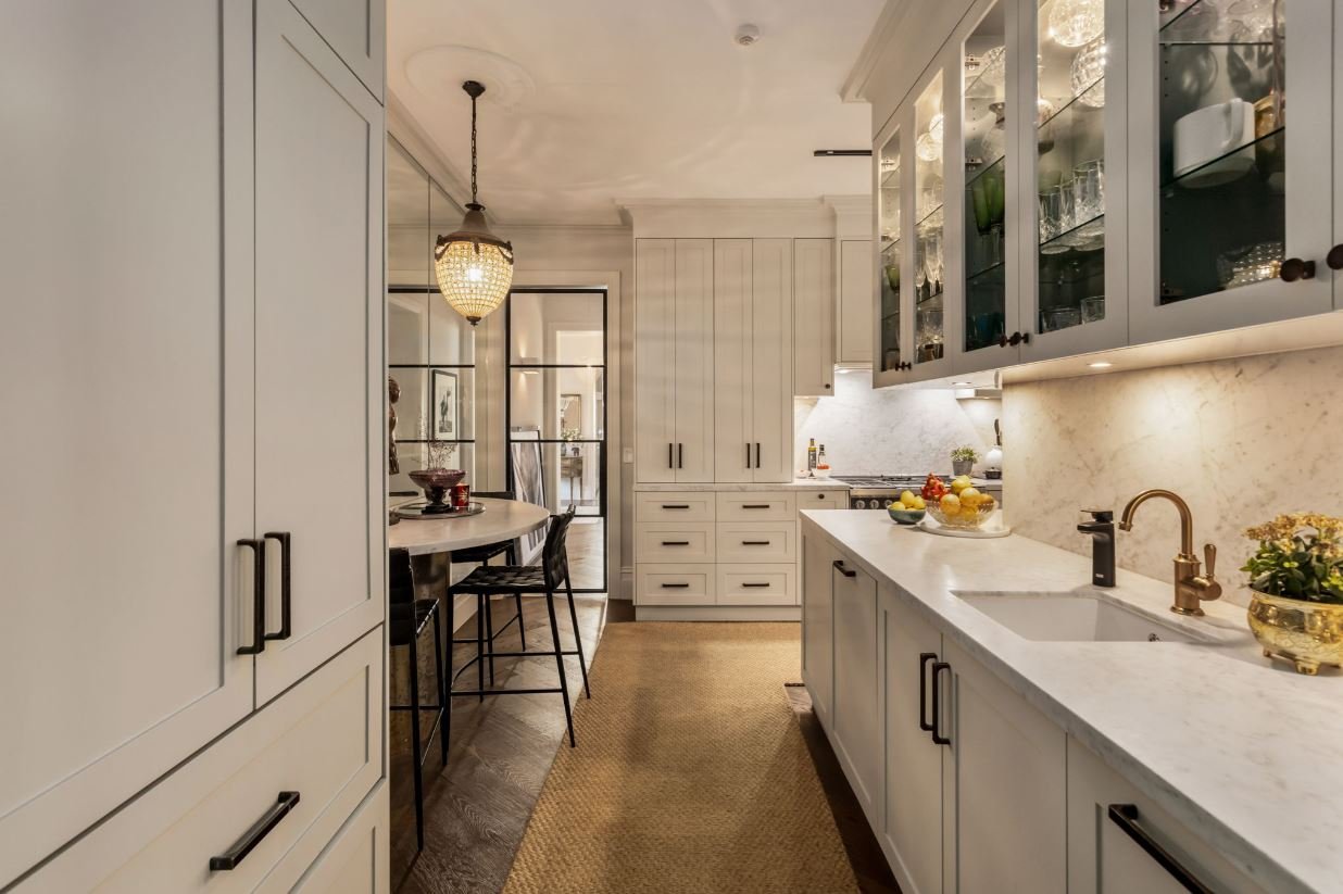 Modern kitchen with white cabinetry, marble countertops, glass-front cabinets, and a dining area with black chairs and a round table with a chandelier.
