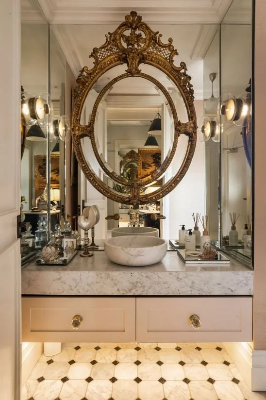 Luxurious bathroom featuring a marble countertop, a white vessel sink, ornate gold-framed mirror, and elegant decor items including soap dispensers and reed diffusers. The space has a vintage design with warm lighting and patterned flooring.