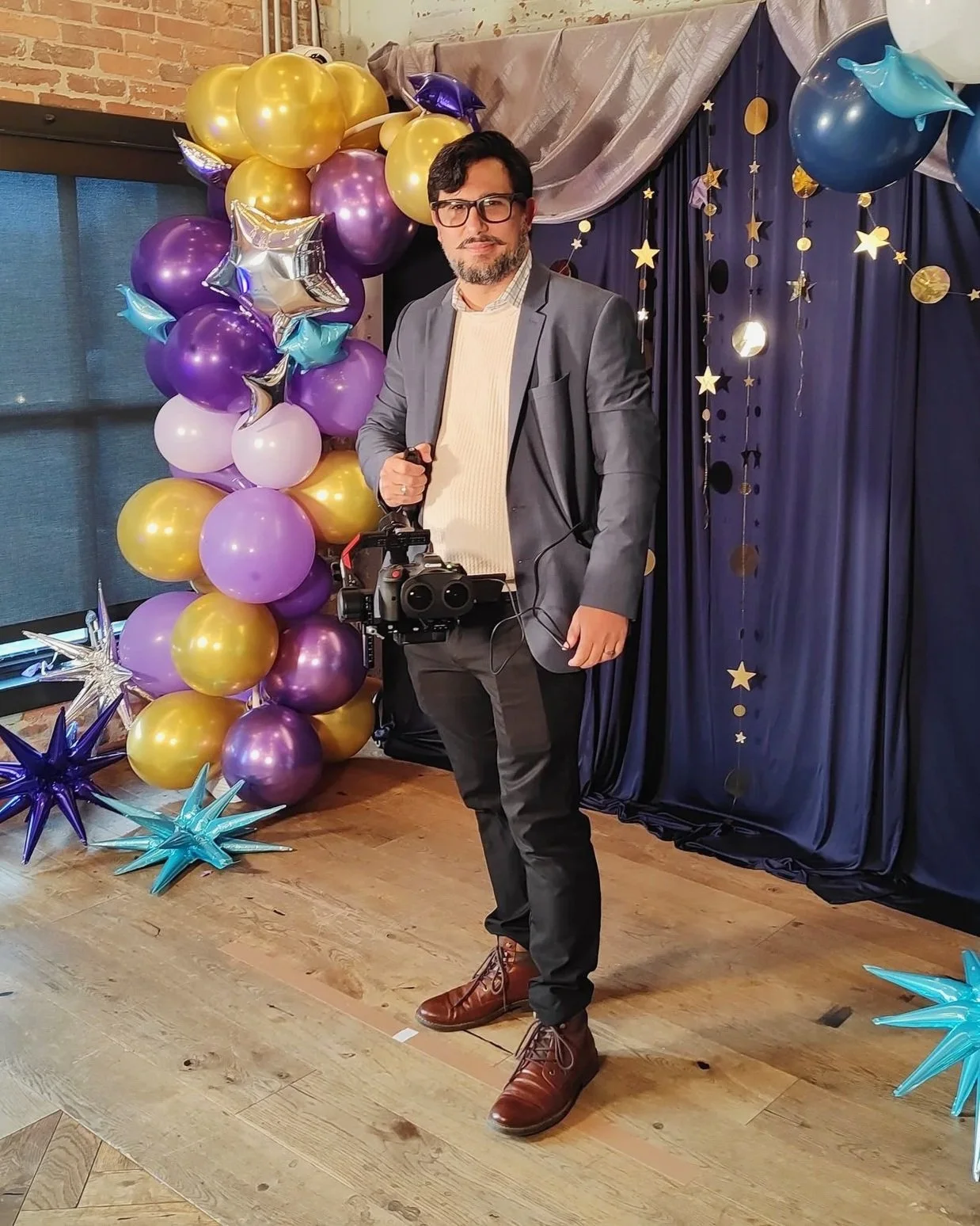 A man in a gray blazer, light sweater, black pants, and brown shoes holding a camera, standing in front of a backdrop with hanging stars and planets, surrounded by gold, purple, pink, blue, and silver balloons and star-shaped balloons on the floor, in a decorated indoor setting.