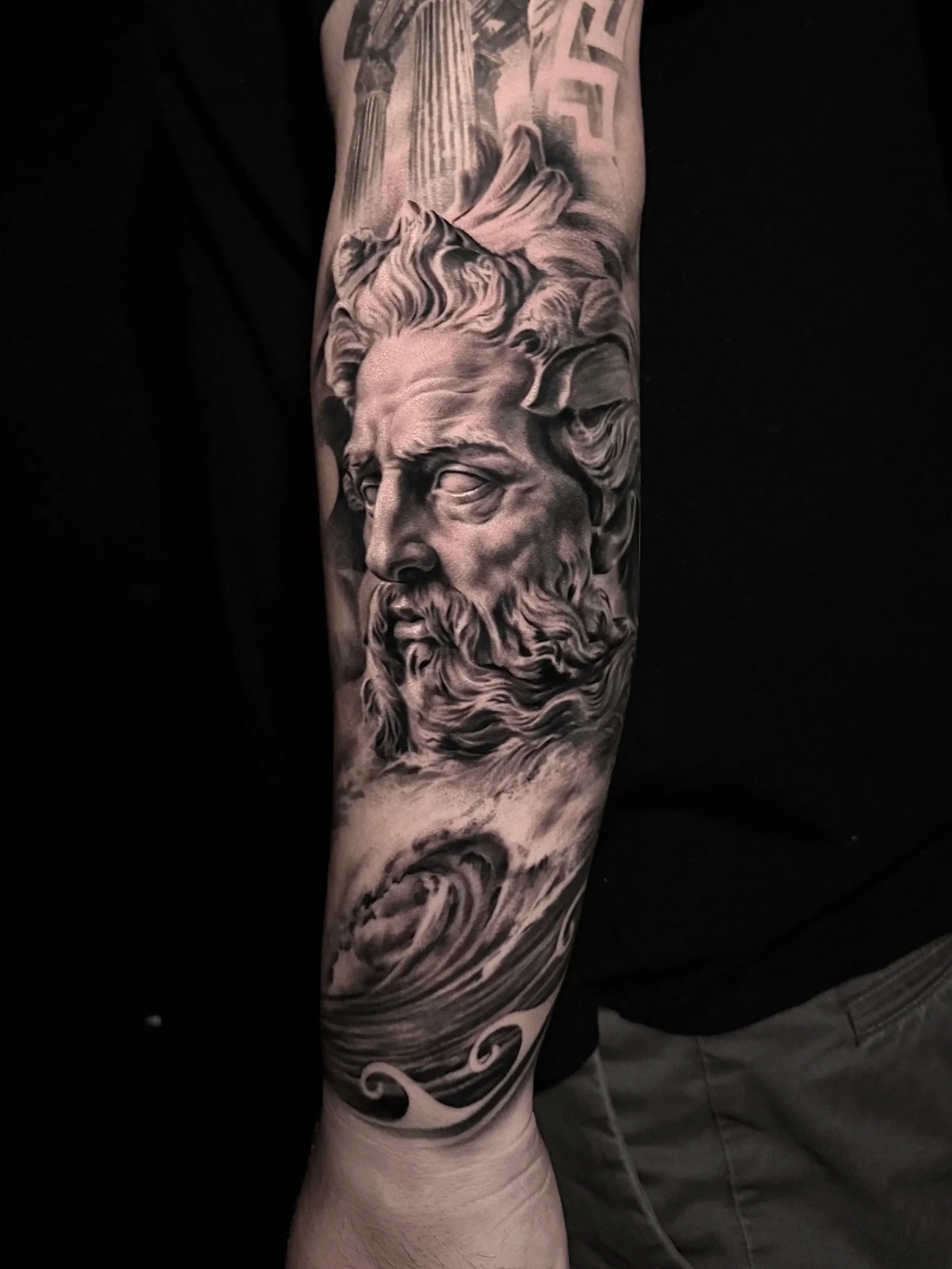 Black and gray realistic tattoo on a person's forearm depicting Poseidon with a stormy sea beneath him and ancient ruins in the background. By Jhon Gutti. In Los Angeles Area, at Outer Limits tattoo shop.