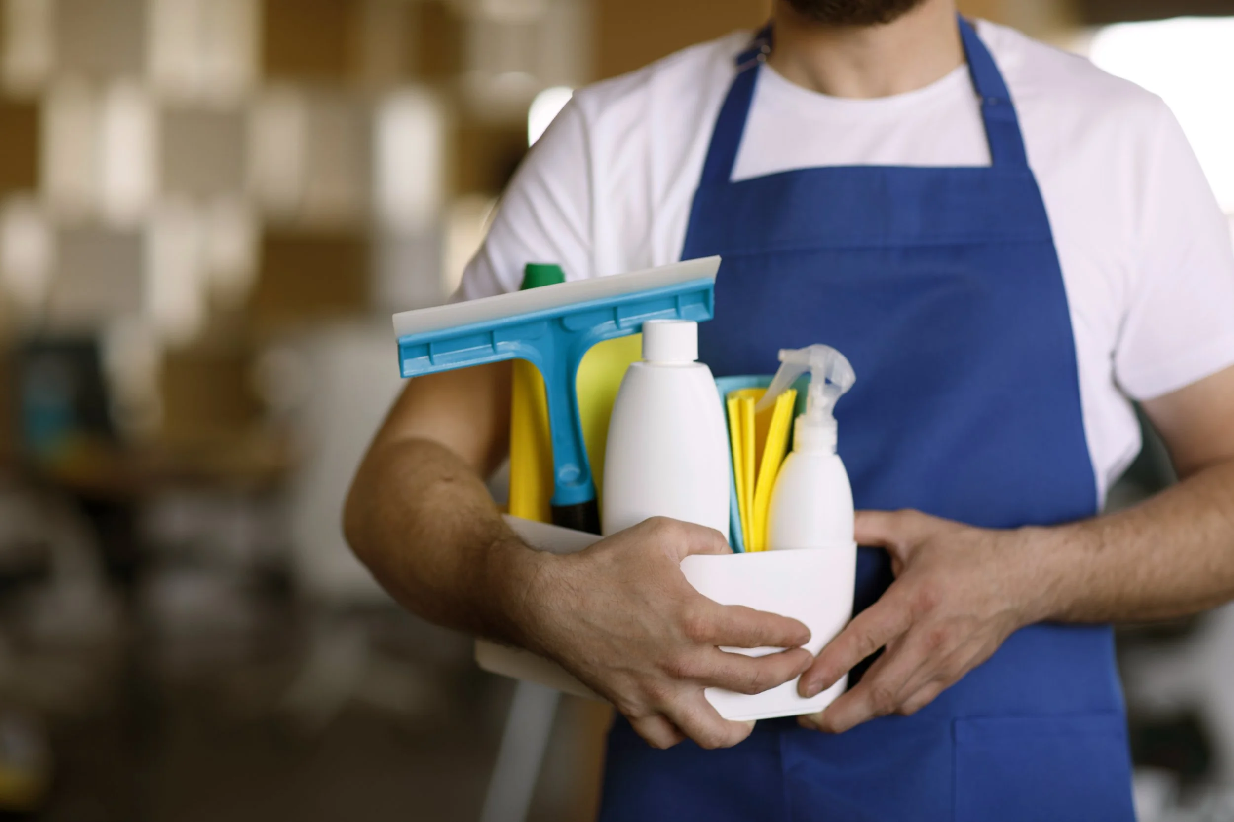 Person wearing a white shirt and blue apron holding a cleaning caddy with various cleaning supplies, including spray bottles, a mop, and cleaning cloths, in a store or cleaning space.