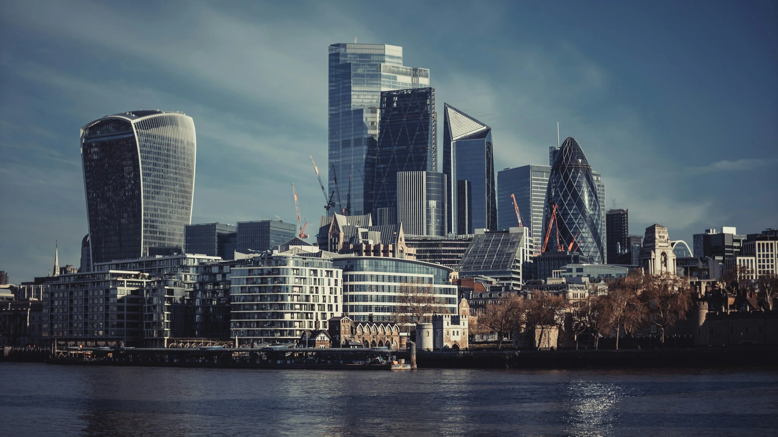 Skyscrapers and modern buildings in London's city skyline viewed from the river under a cloudy sky.