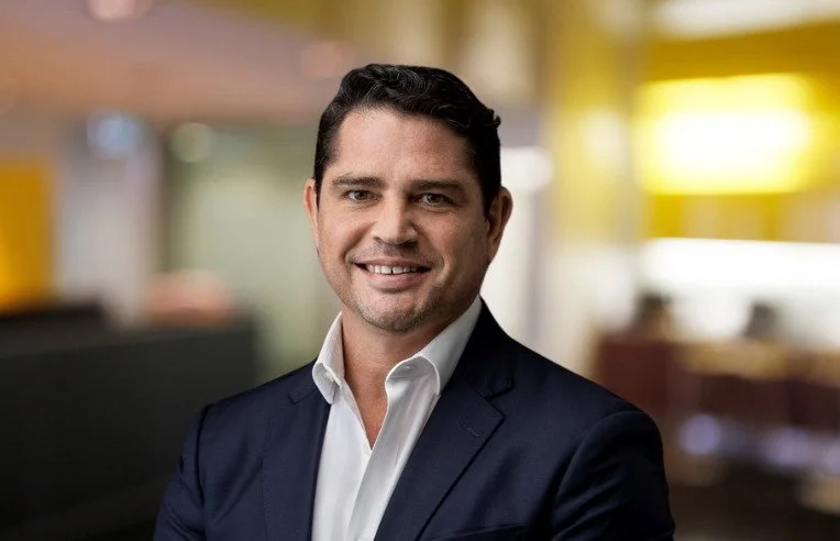 A man in a navy suit and white shirt smiling in an indoor setting with blurred background.