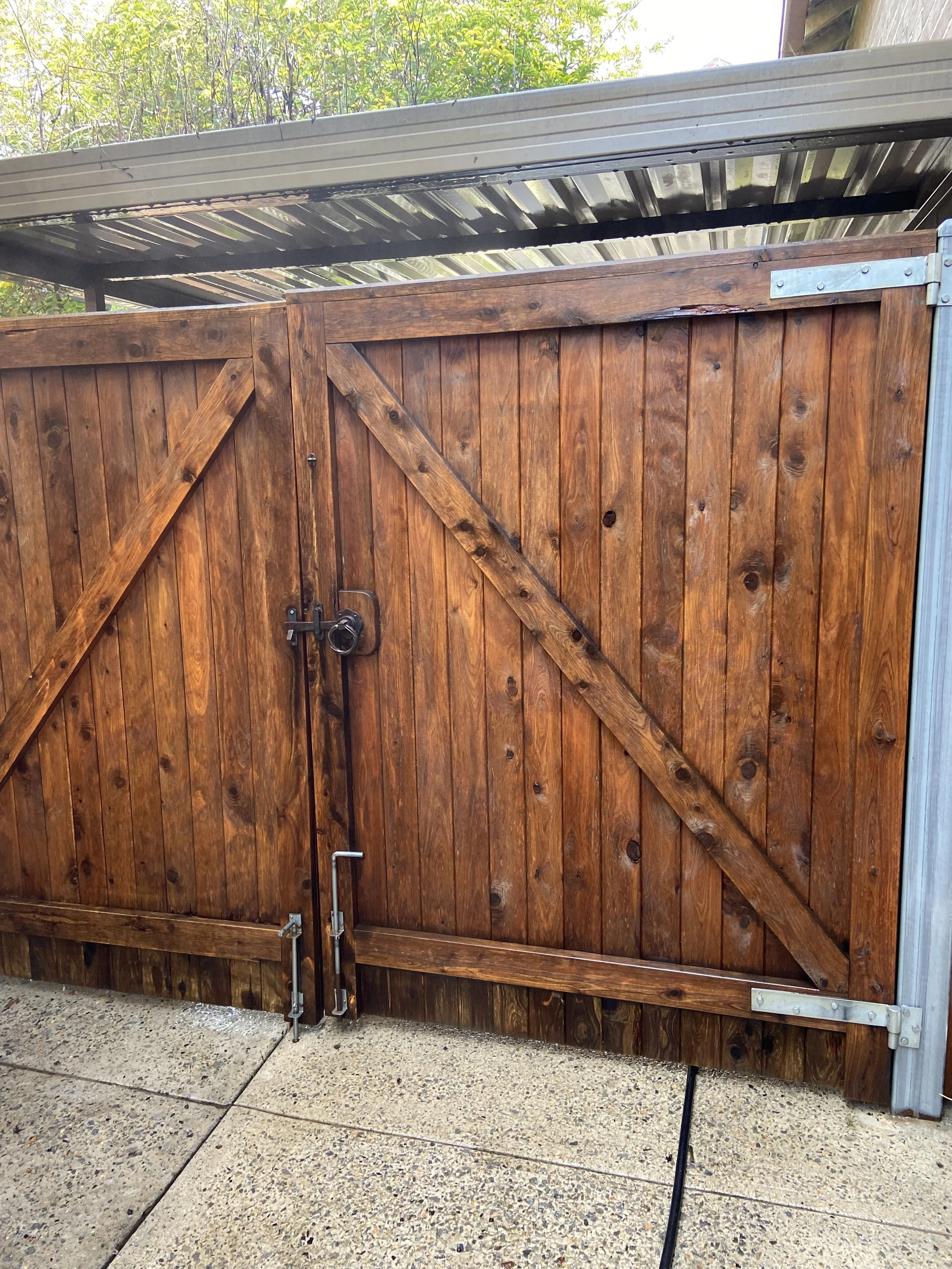 Wooden gate with metal hinges and lock, set on a concrete surface with a metal roof and greenery overhead.