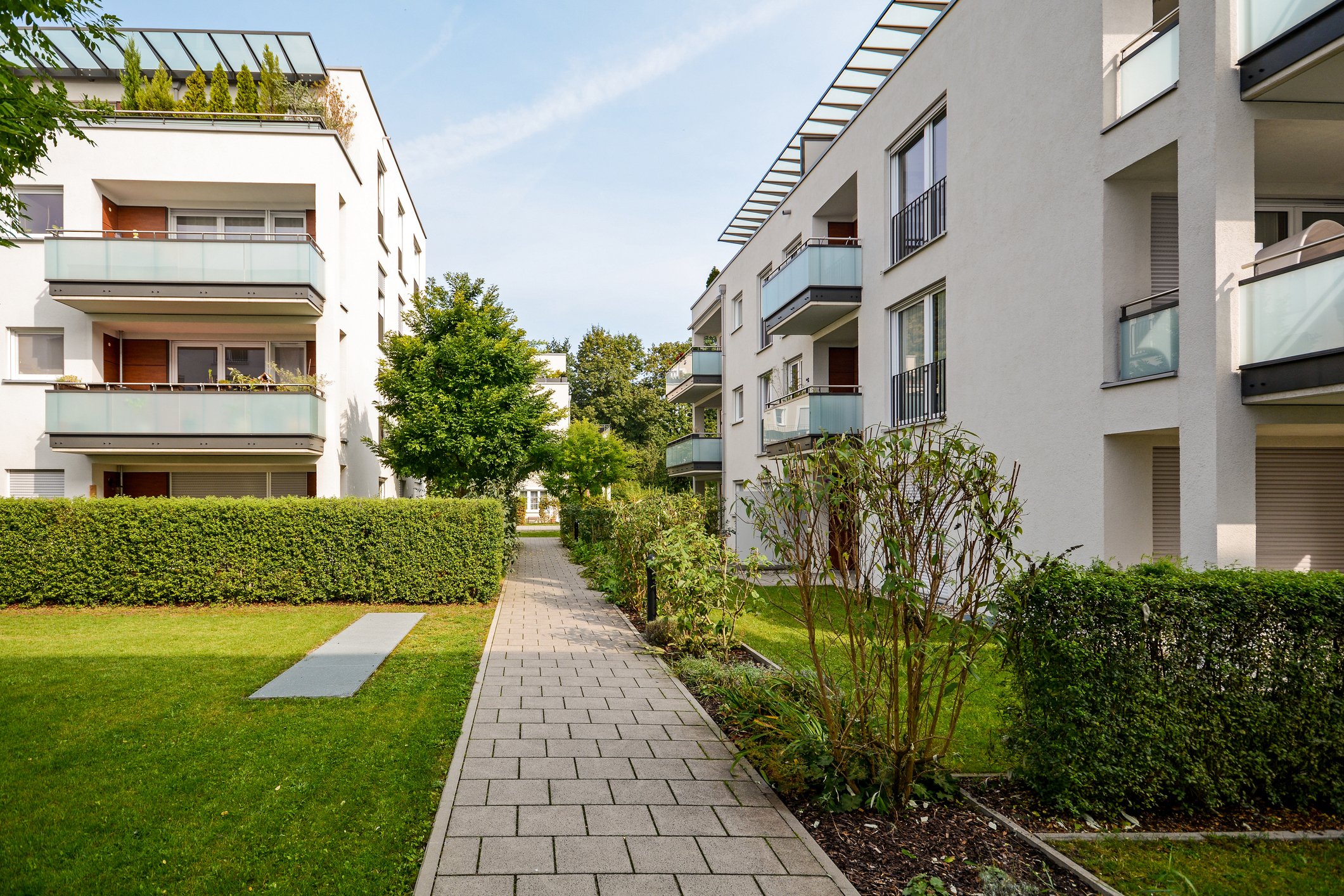 A paved sidewalk in an apartment strata complex courtyard, with green grass, shrubs, and trees on either side, and white multi-story buildings with balconies and sliding glass doors.