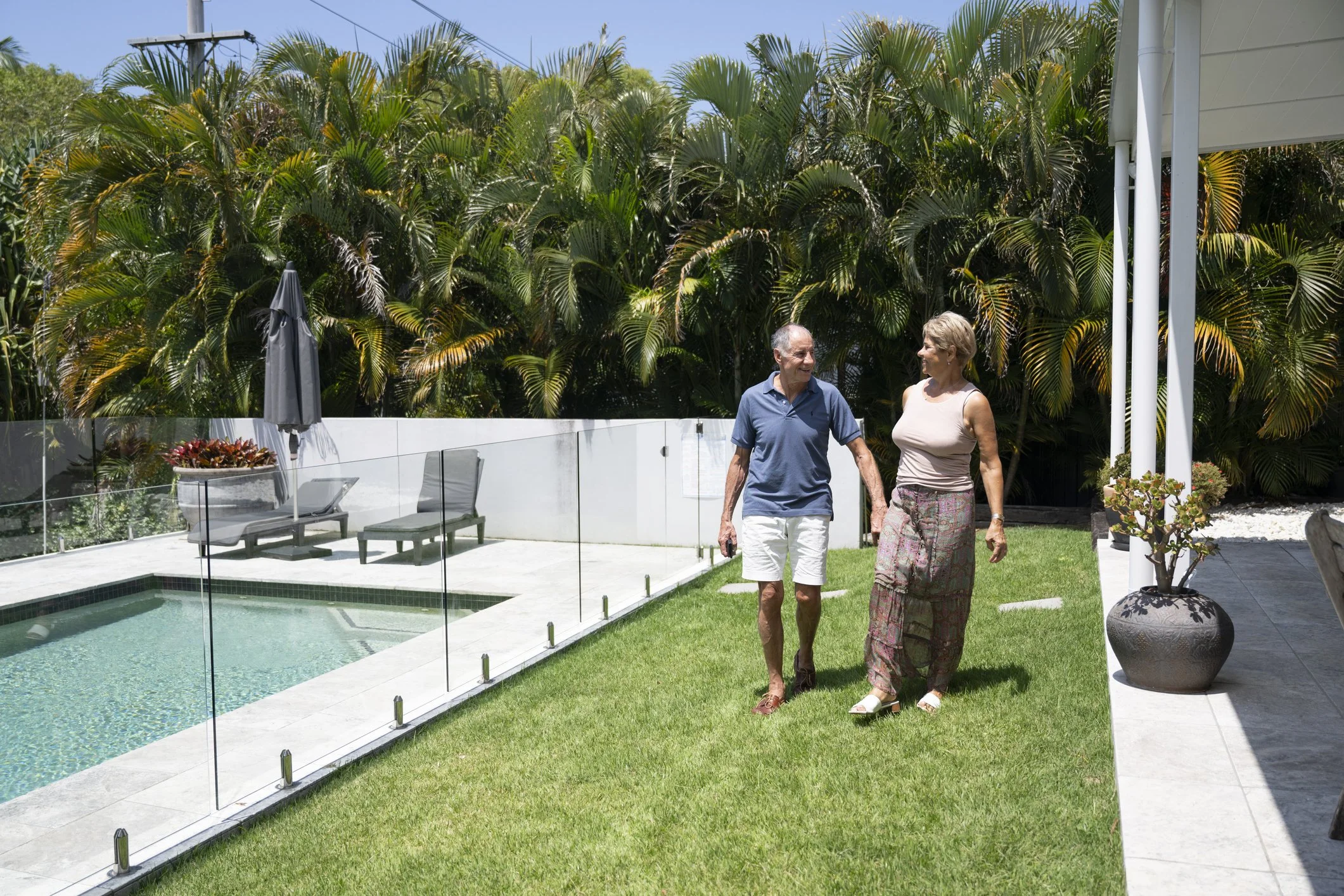 Older man and woman walking and talking by a backyard pool surrounded by lush palm trees and tropical plants on a sunny day.