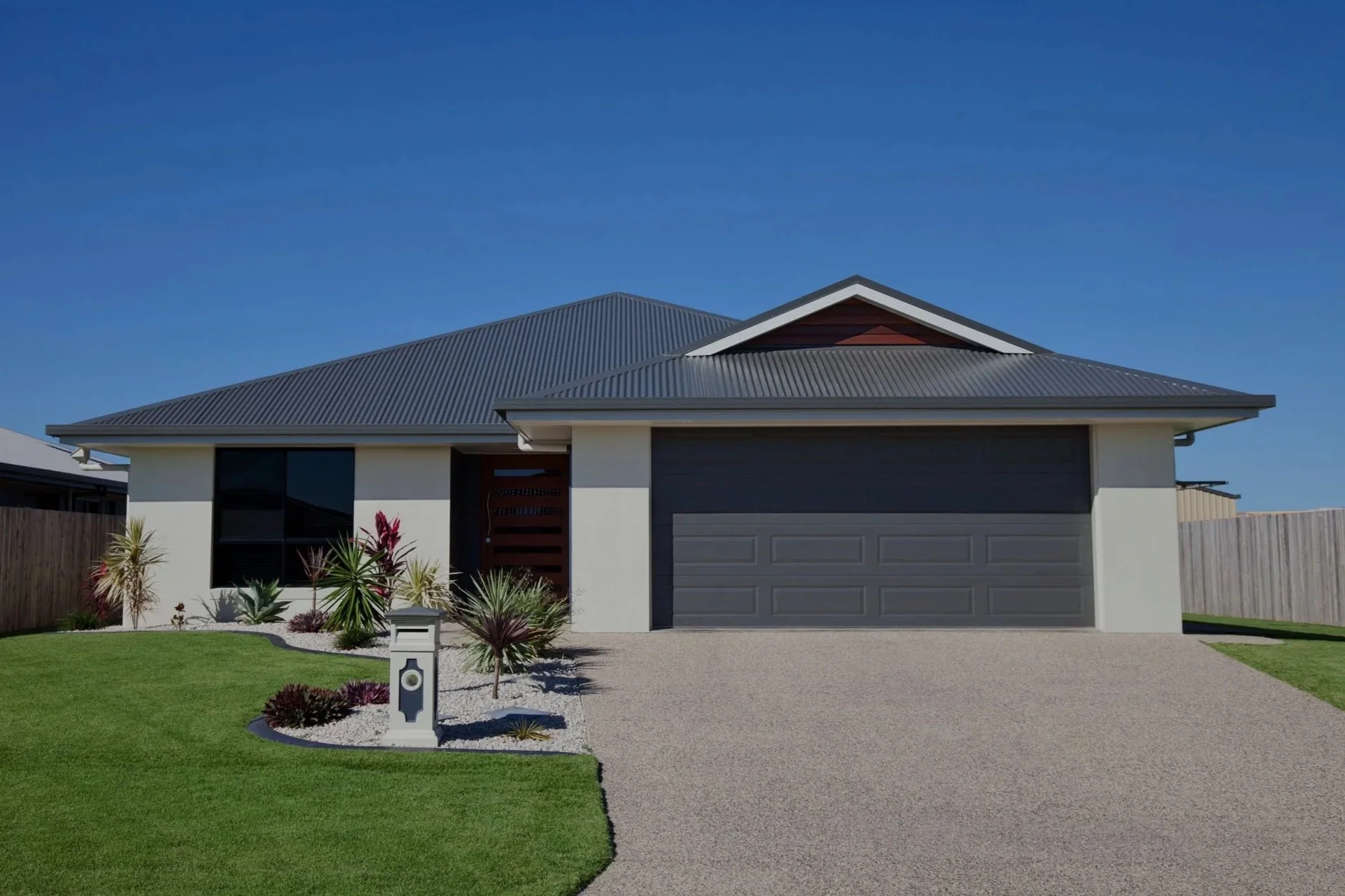 Front view of a modern single-story house with a grey garage door, white exterior walls, a grey metal roof, and a landscaped front yard with green grass, decorative plants, and a white mailbox, against a clear blue sky.