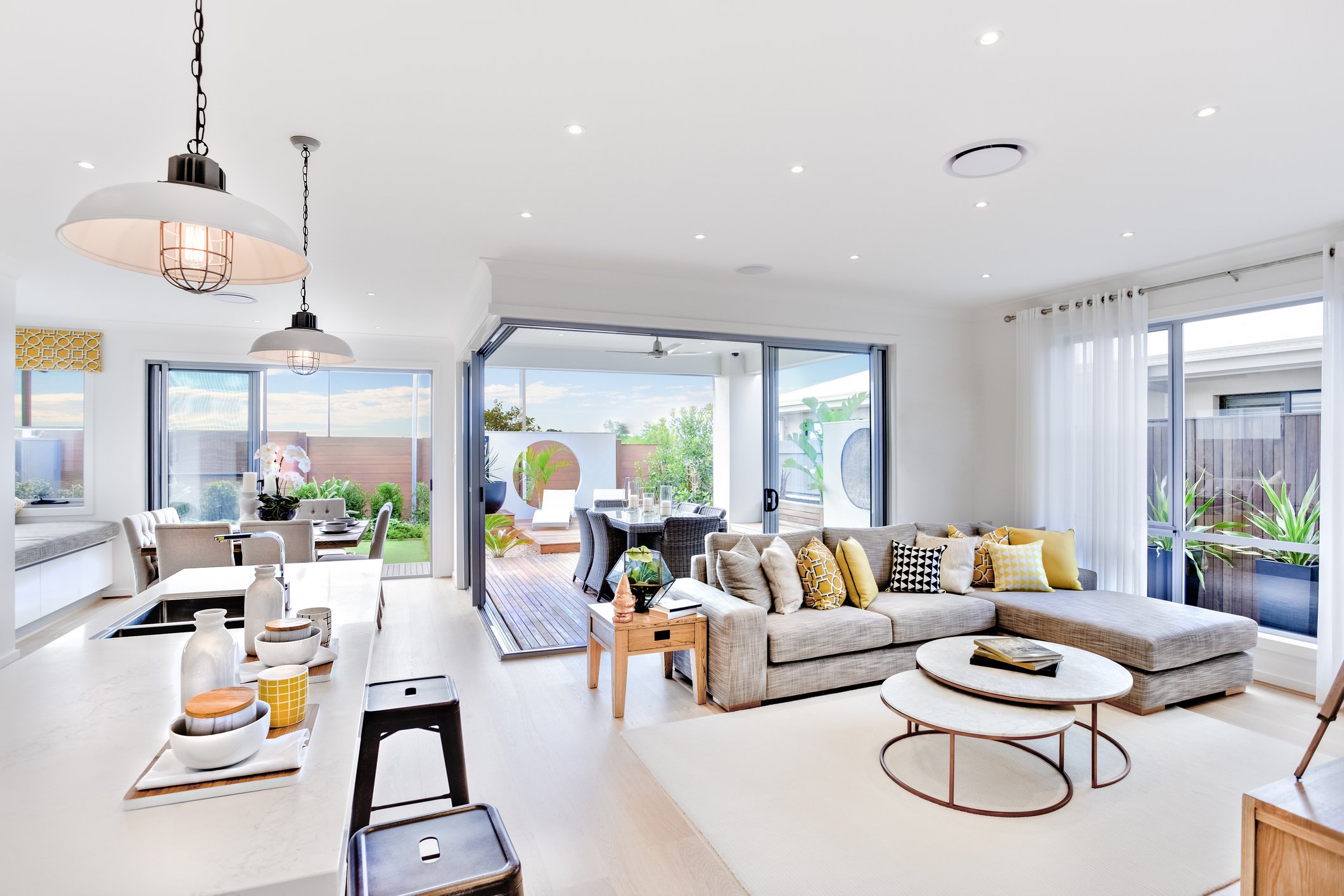 Bright display home living room with large windows, open patio with outdoor dining, beige sectional sofa with colourful pillows, and modern decor.