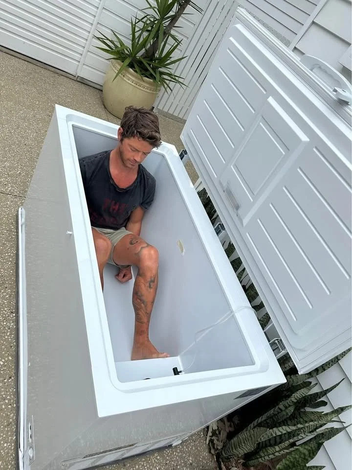 A man sitting inside an empty ice chest on an outdoor patio, with white fencing and potted plants around.