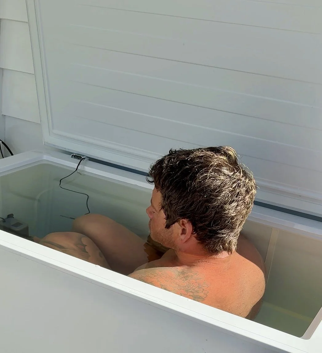 A man with tattooed shoulder relaxing in a white outdoor ice bath tub with a cover. The tub is filled with water and the cover is open.