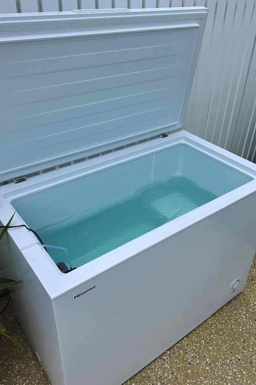 A white Hisense chest freezer with the lid open, filled with water, located outdoors next to a wall with vertical siding.