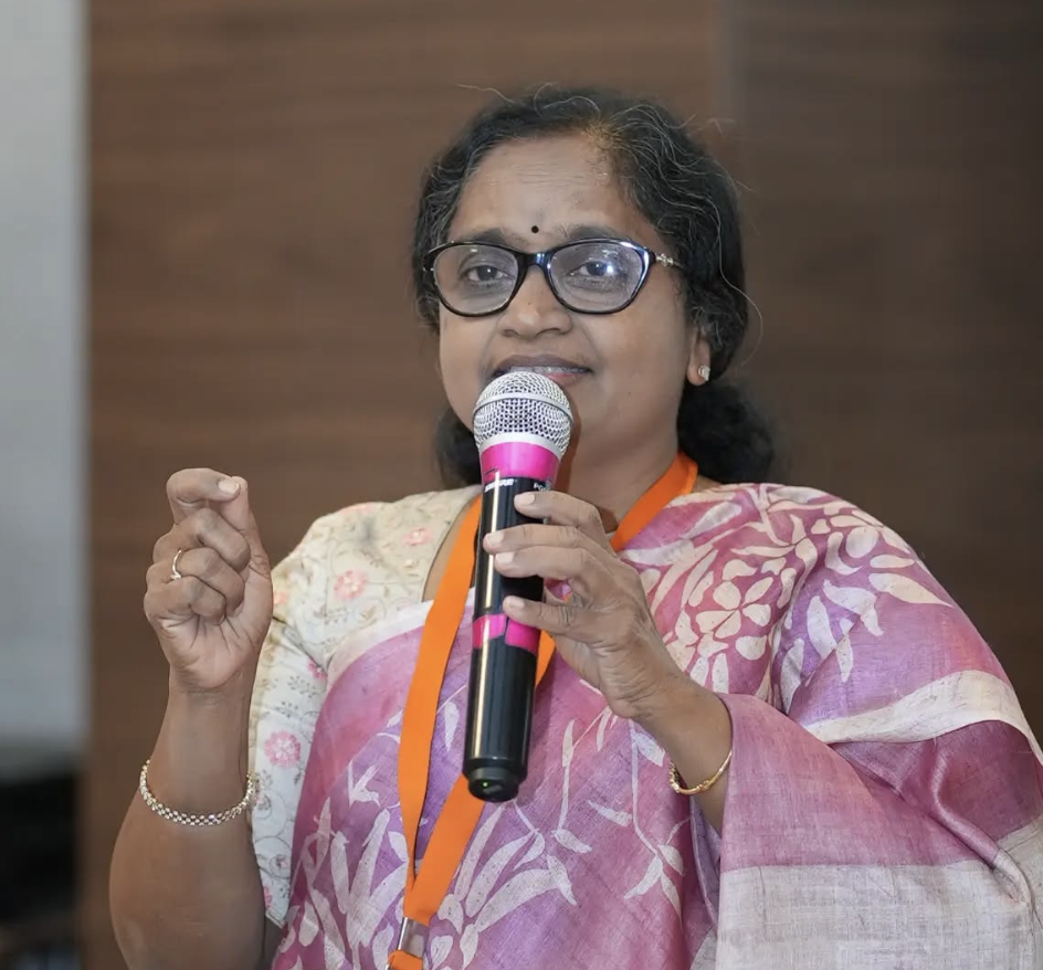 A woman with glasses and a pink saree speaking into a microphone during an event.