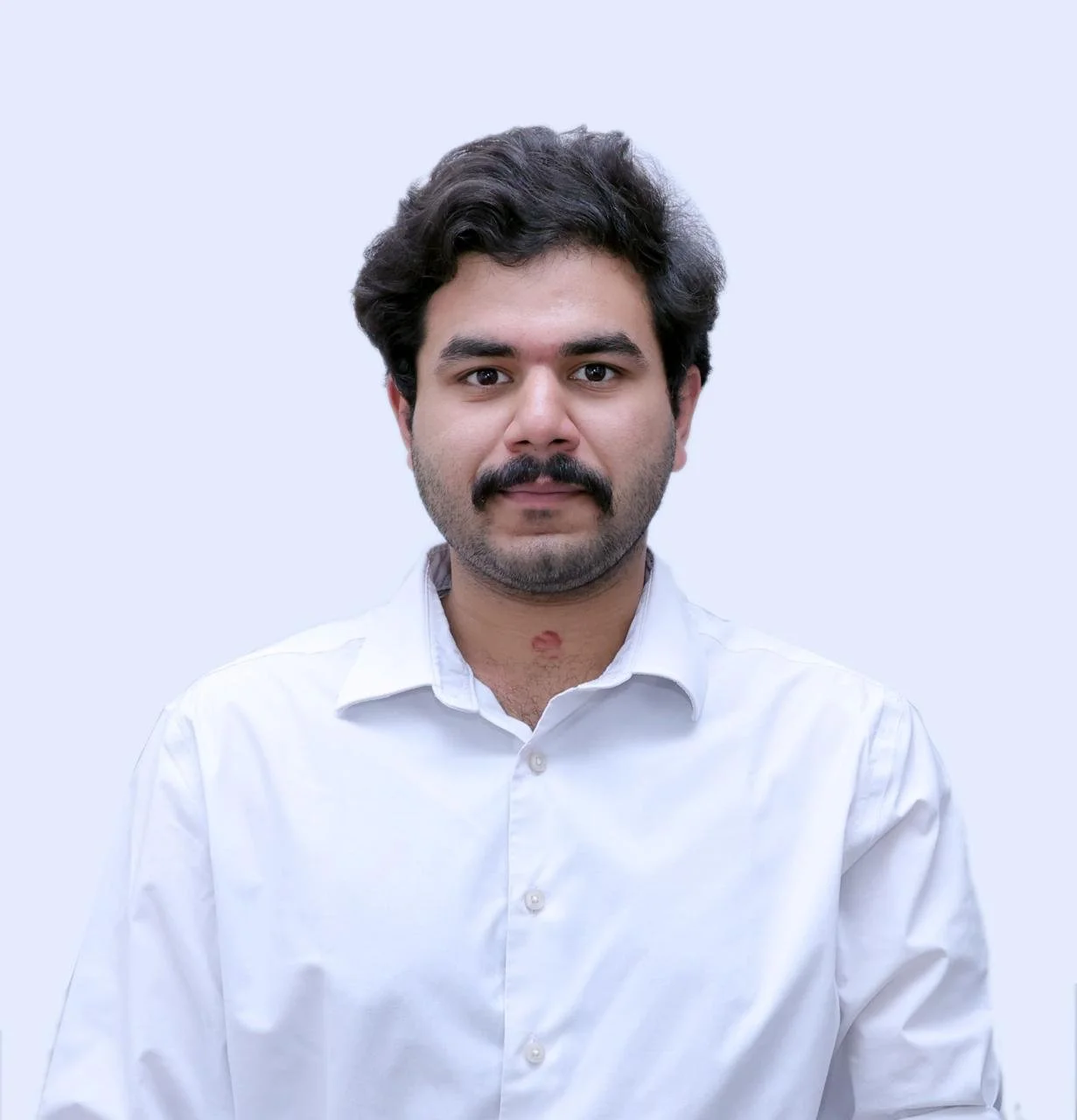 A man with dark hair and a mustache wearing a white button-up shirt, standing against a light gray background.