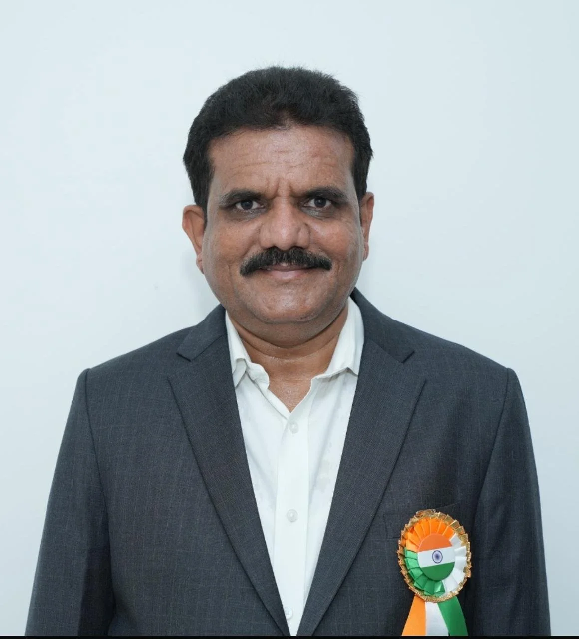 Man with dark hair and mustache wearing a formal dark gray suit and white shirt, standing against a plain white background. He has a ribbon badge with Indian flag colors on his left chest.