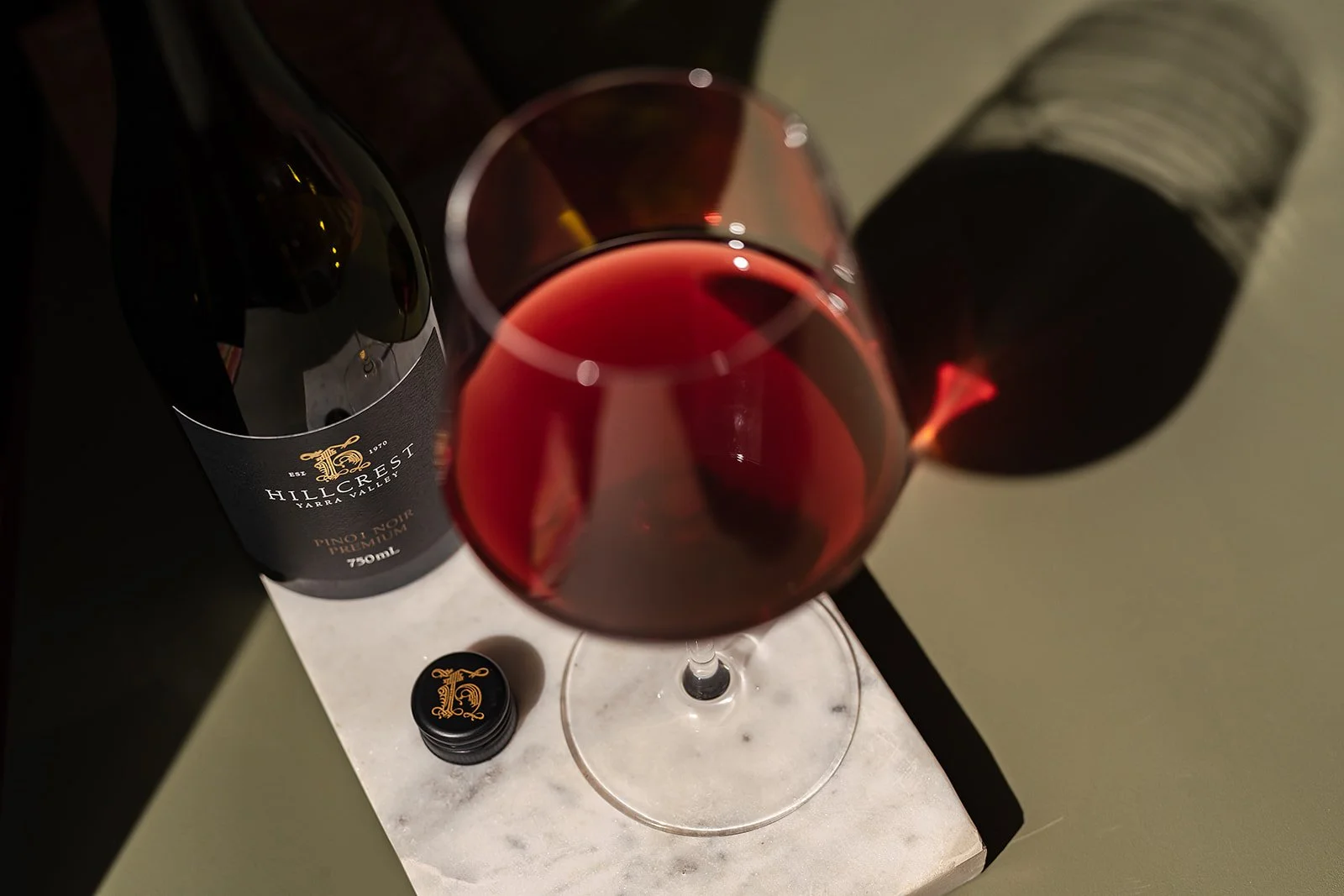 A glass of red wine on a white marble block with a bottle of Hillcrest Zarra Valley Primoire wine and a black wine cap nearby.
