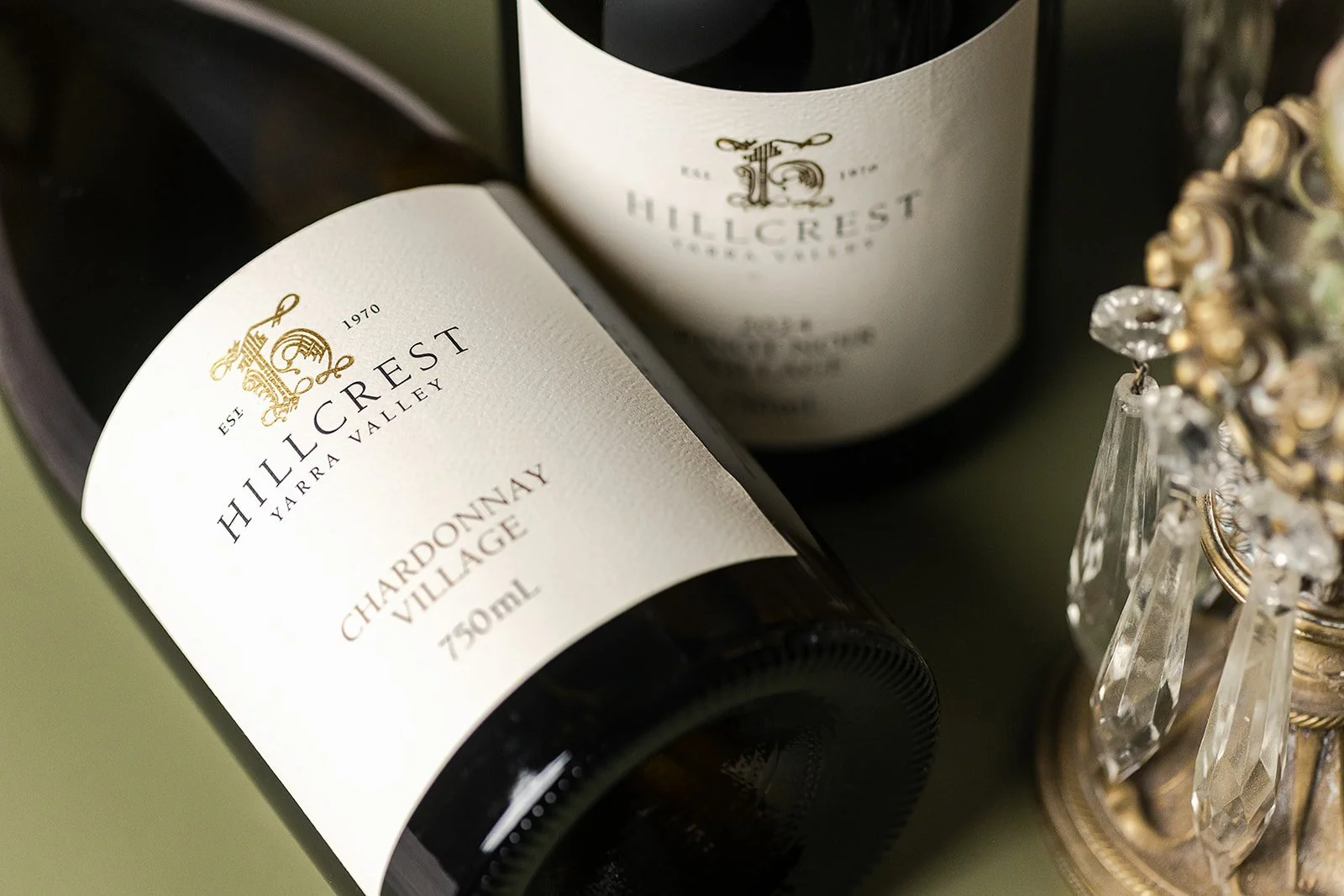 Close-up of two bottles of Hillcrest Yarra Valley Chardonnay Village wine on a table, with a crystal and gold decorative piece nearby.