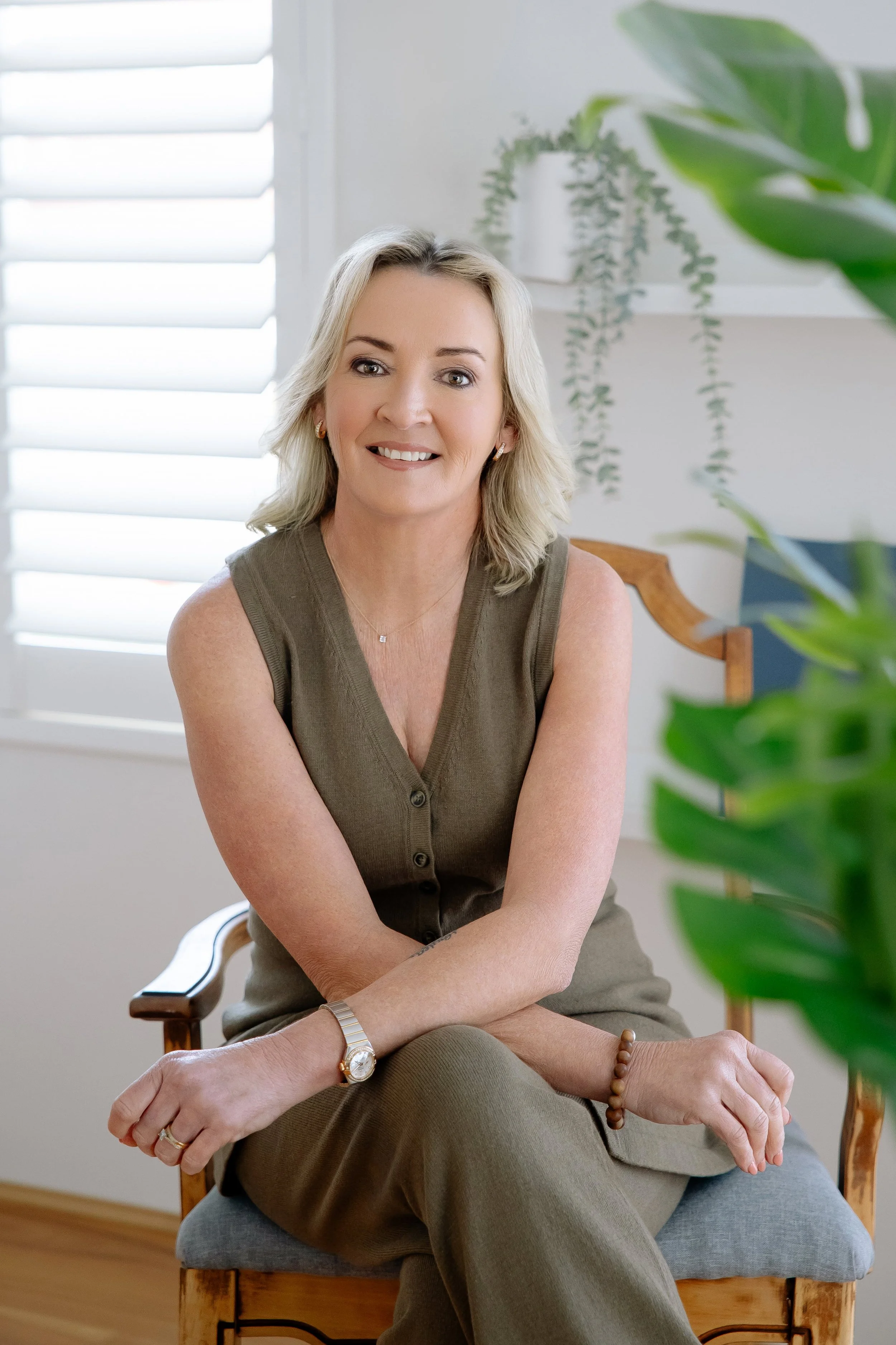 A middle-aged woman with blonde hair, wearing a sleeveless olive green top and matching pants, sitting on a wooden chair inside a well-lit room with white walls and plants in the background.
