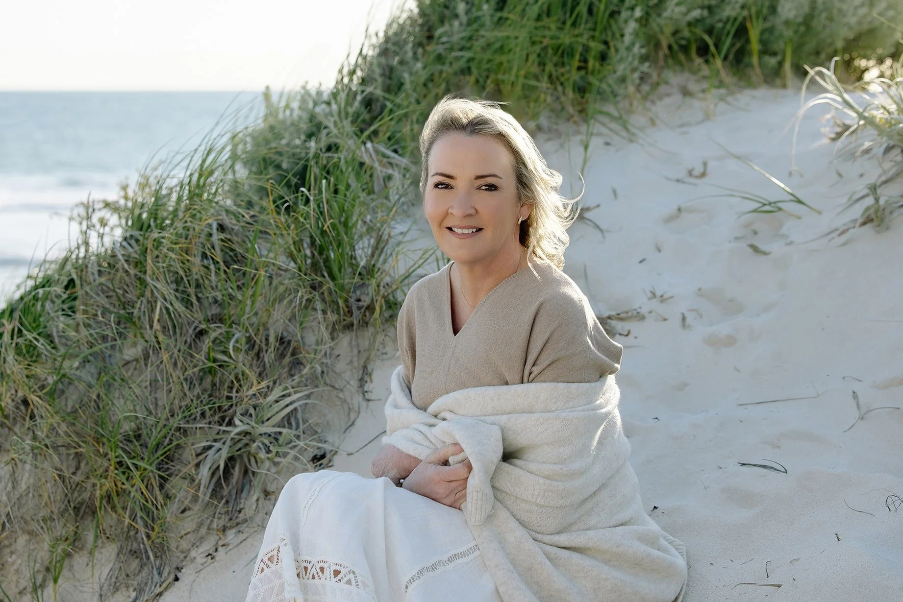 A middle-aged woman sitting on a sandy beach near green beach grass, wearing a beige top and a cream shawl, smiling at the camera during daytime.