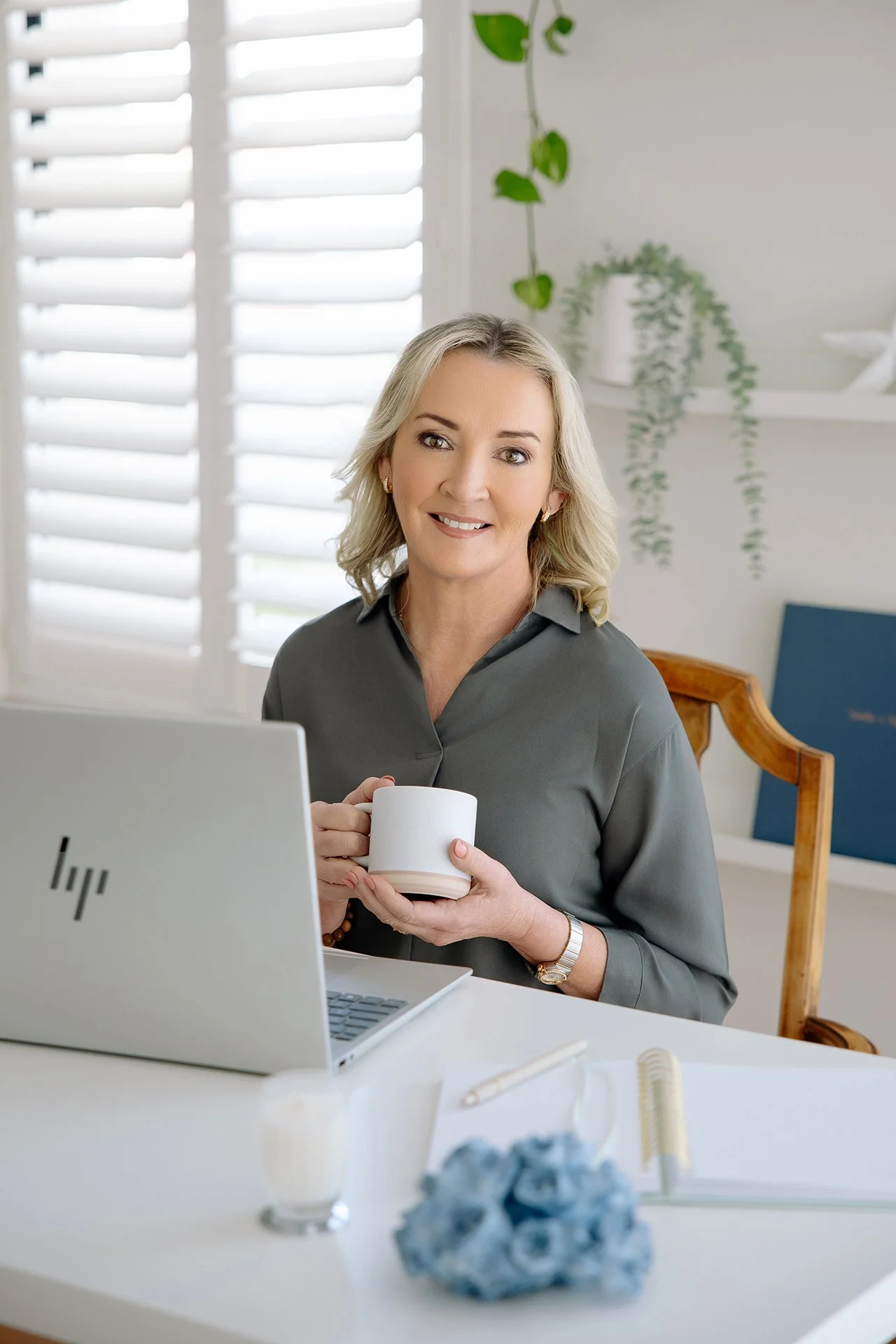 A woman with blonde hair and a gray blouse sitting at a desk in front of a laptop, holding a white mug, with a bright window and green plants in the background.