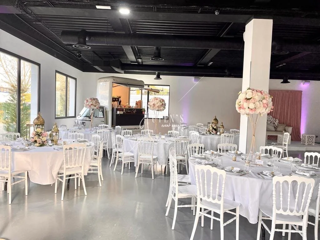 Salle de réception élégante avec tables rondes et longues, décorée de fleurs roses et blanches, fenêtre laissant entrer la lumière naturelle, mobilier blanc et ambiance sophistiquée pour un événement spécial.